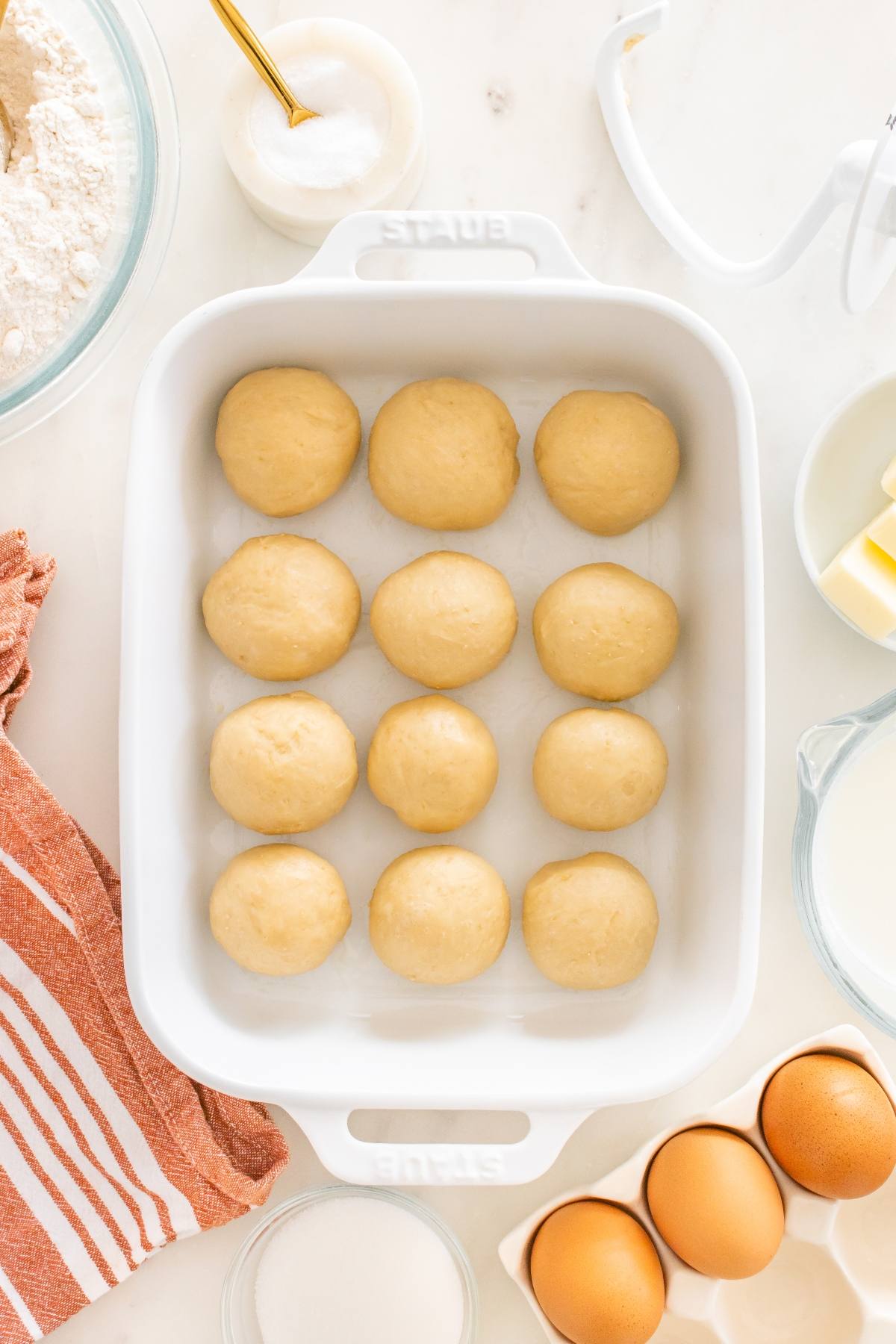 Twelve dough balls in a white baking dish, surrounded by baking ingredients on a white counter.