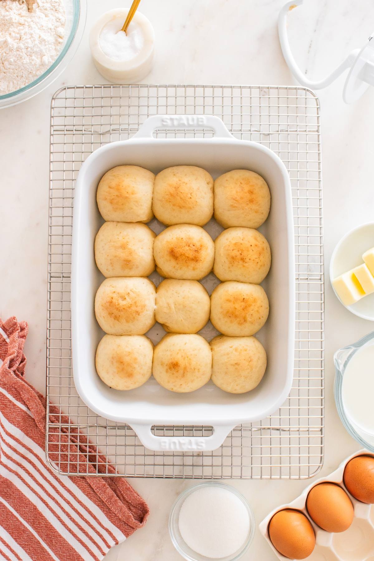 A white baking dish with twelve golden brown dinner rolls on a cooling rack, surrounded by baking ingredients.