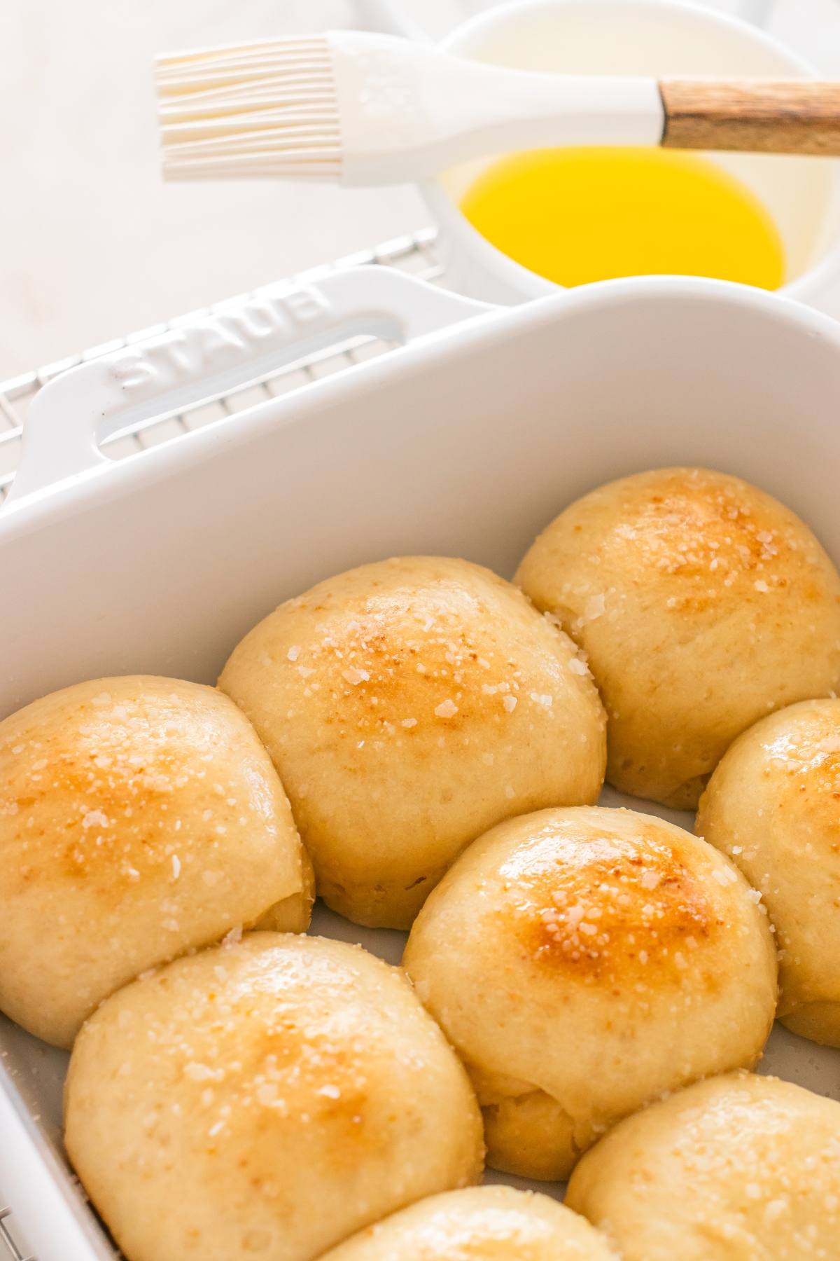 A baking dish of golden dinner rolls topped with coarse salt, with a bowl of melted butter and pastry brush nearby.