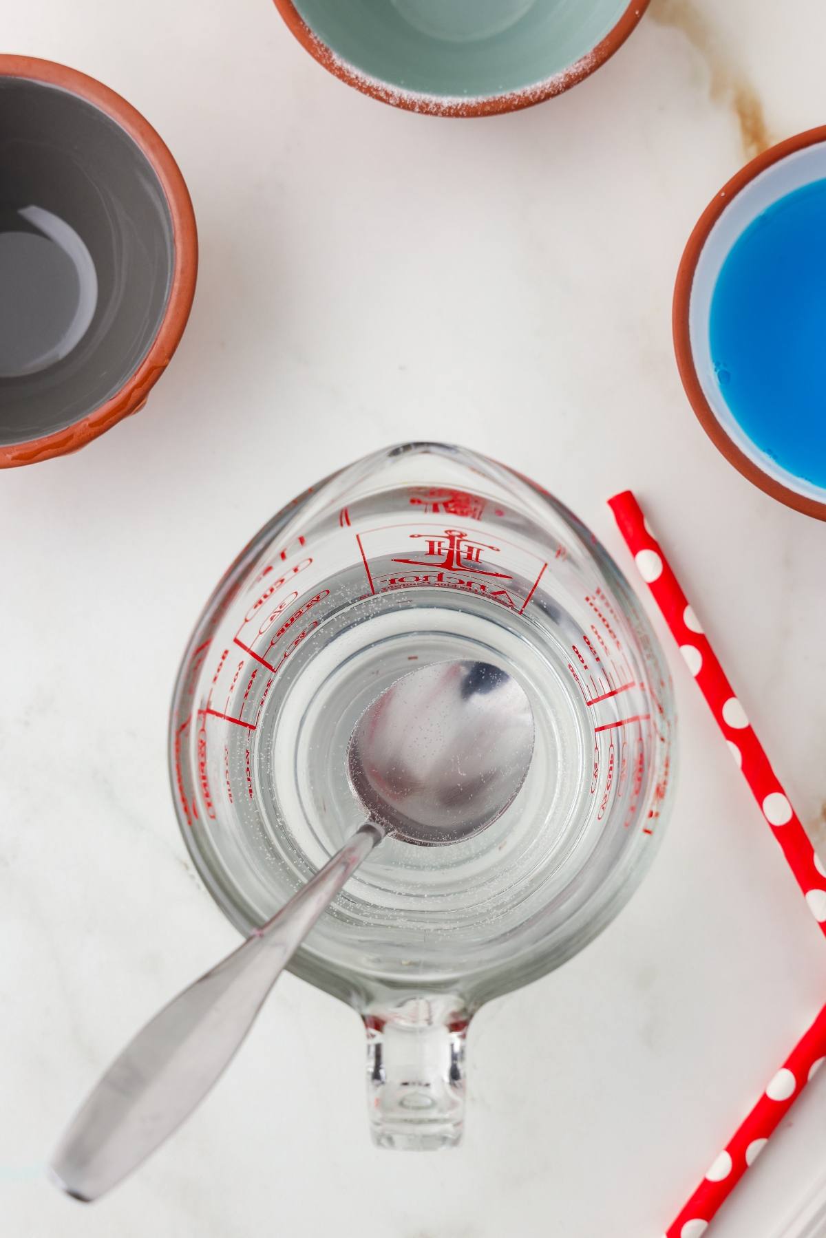 A measuring cup with clear liquid and a spoon, surrounded by bowls and red polka dot straws.