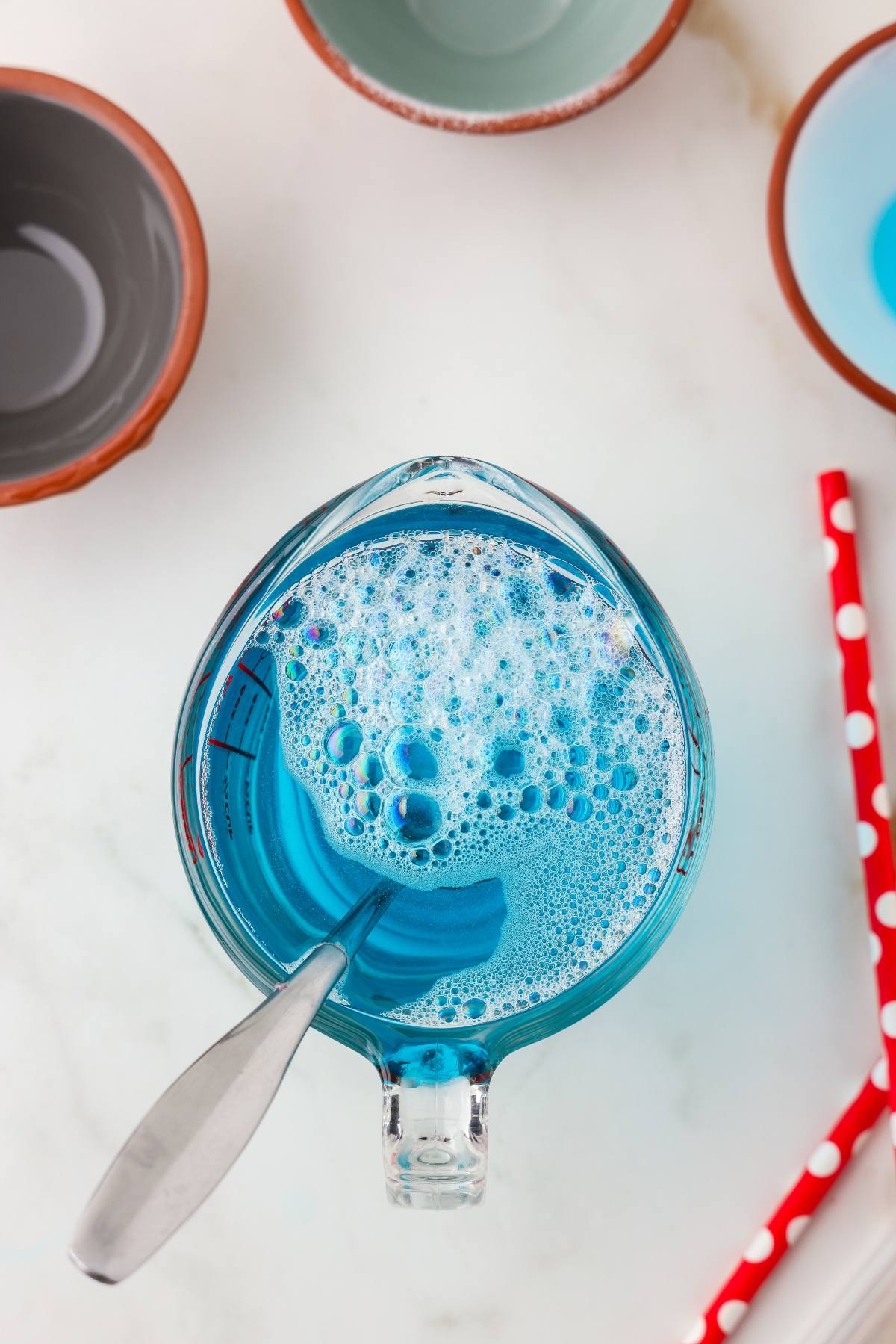 A glass measuring cup with blue liquid and bubbles, a spoon inside, surrounded by bowls and red straws.