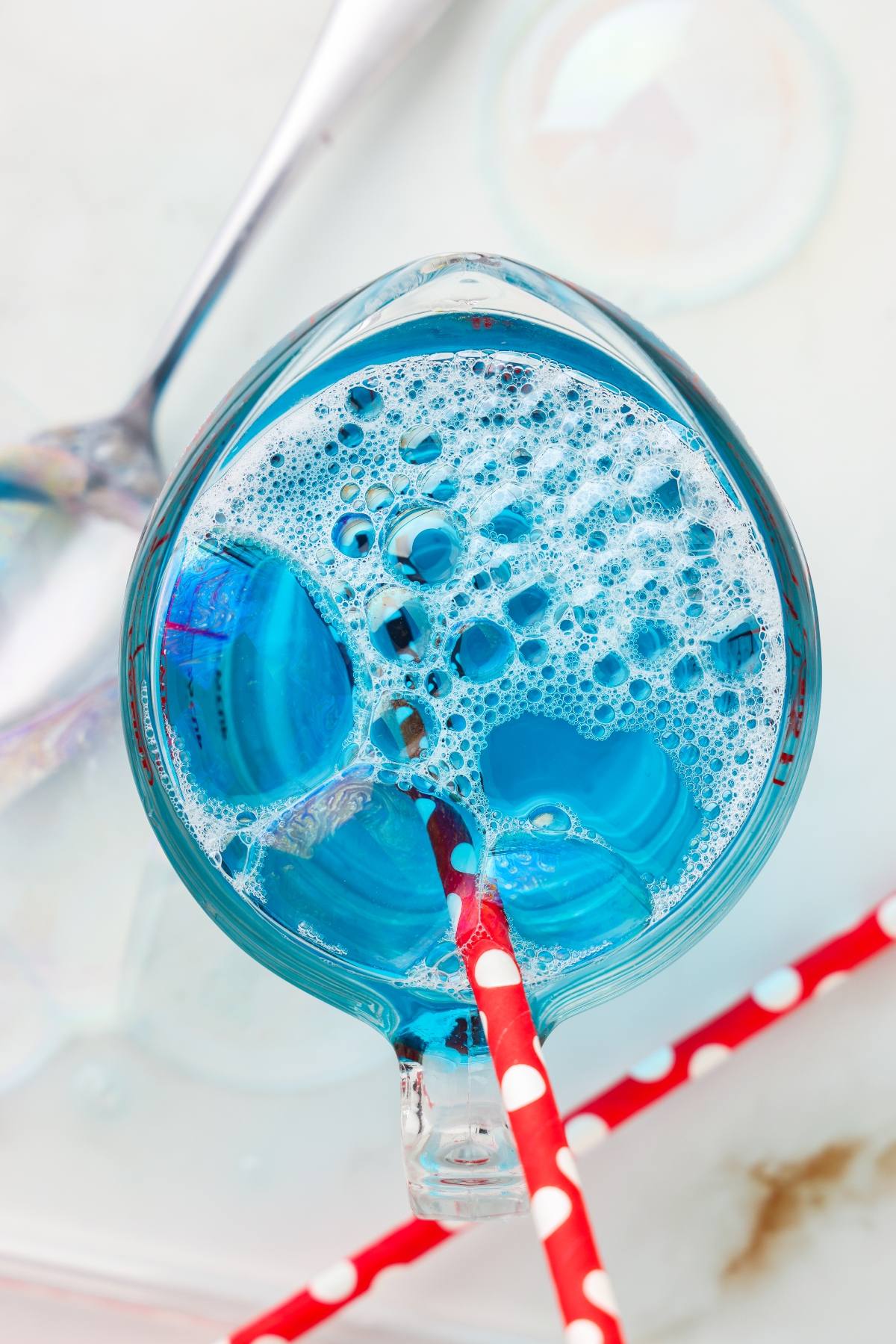 A glass cup with blue bubbly liquid and a red polka-dot straw, viewed from above.