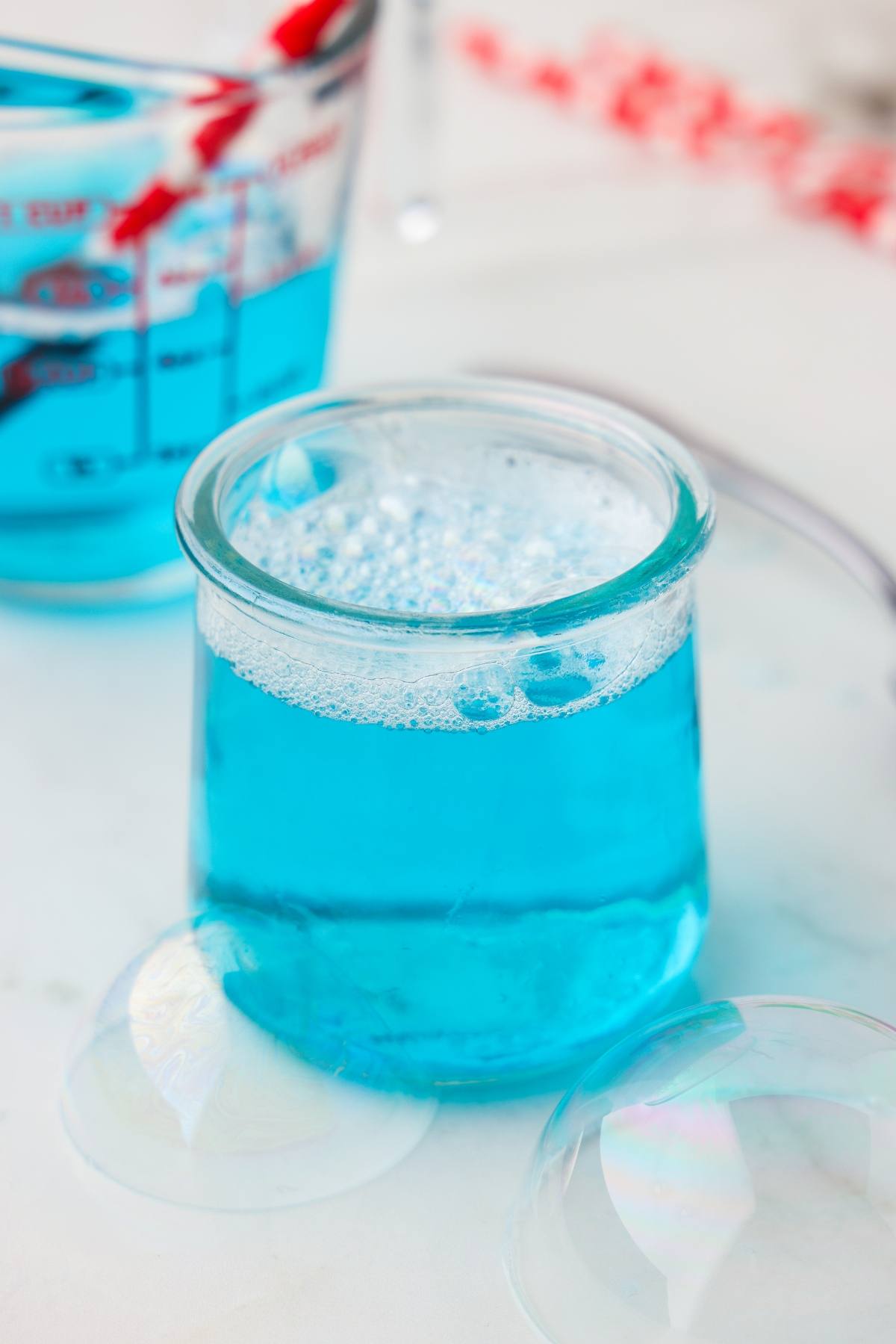 A glass jar filled with blue liquid and bubbles, with a measuring cup of blue liquid in the background.