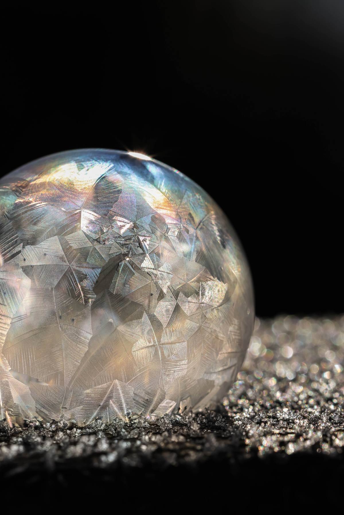 A frozen soap bubble with crystal patterns sits on a sparkling, frosty surface against a dark background.