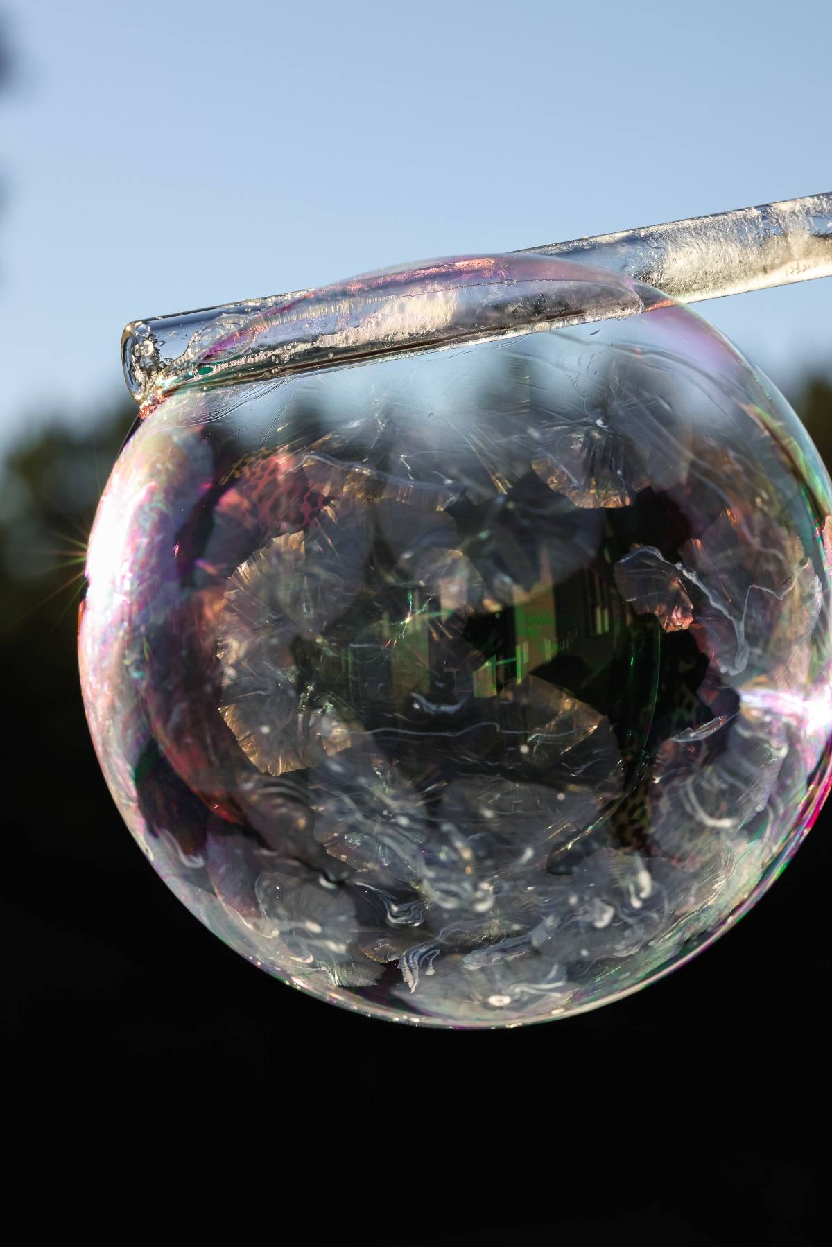 A close-up of a soap bubble forming on a wand, reflecting light and colors against a blurred outdoor background.