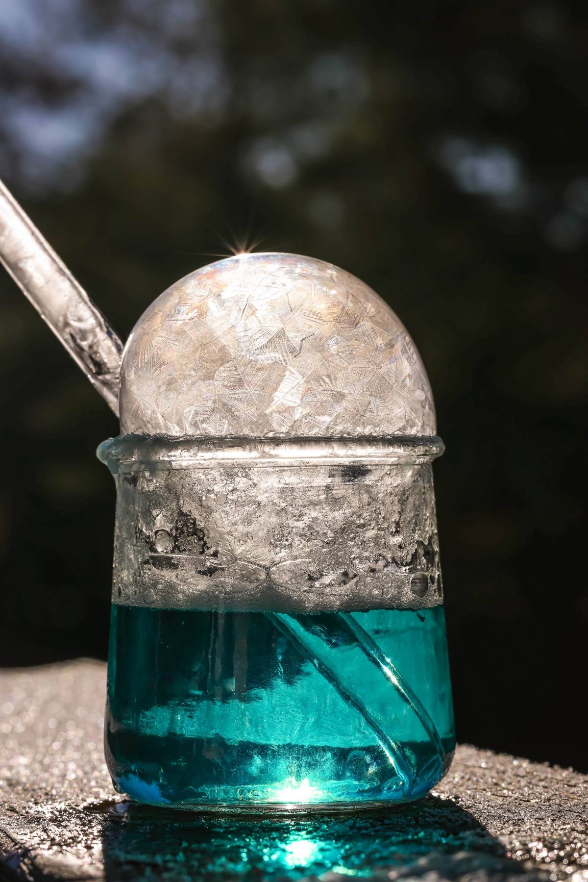 A frozen soap bubble sits on top of a glass of blue liquid, with sunlight sparkling on the ice.