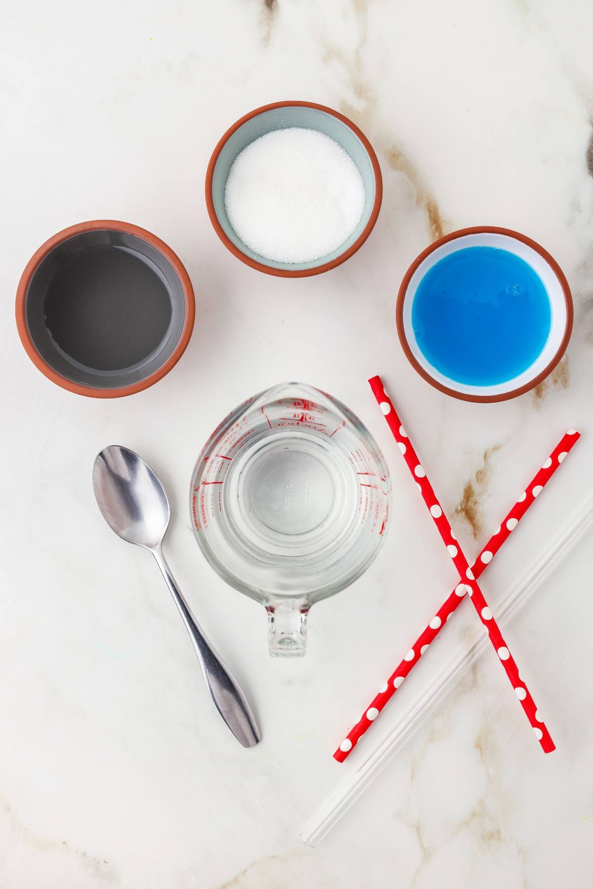 Measuring cup, spoon, sugar, blue liquid, water, two straws, and a bowl on a marble surface.