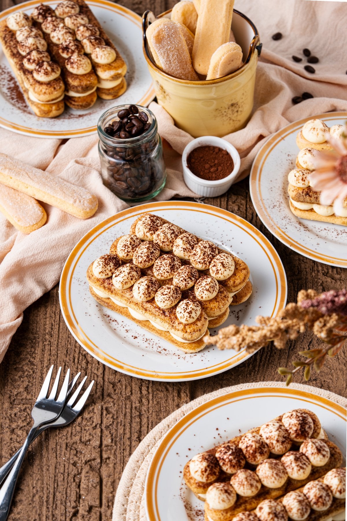Plates of individual tiramisu desserts and ladyfingers rest on a wooden table, surrounded by coffee beans and a dusting of cocoa powder.