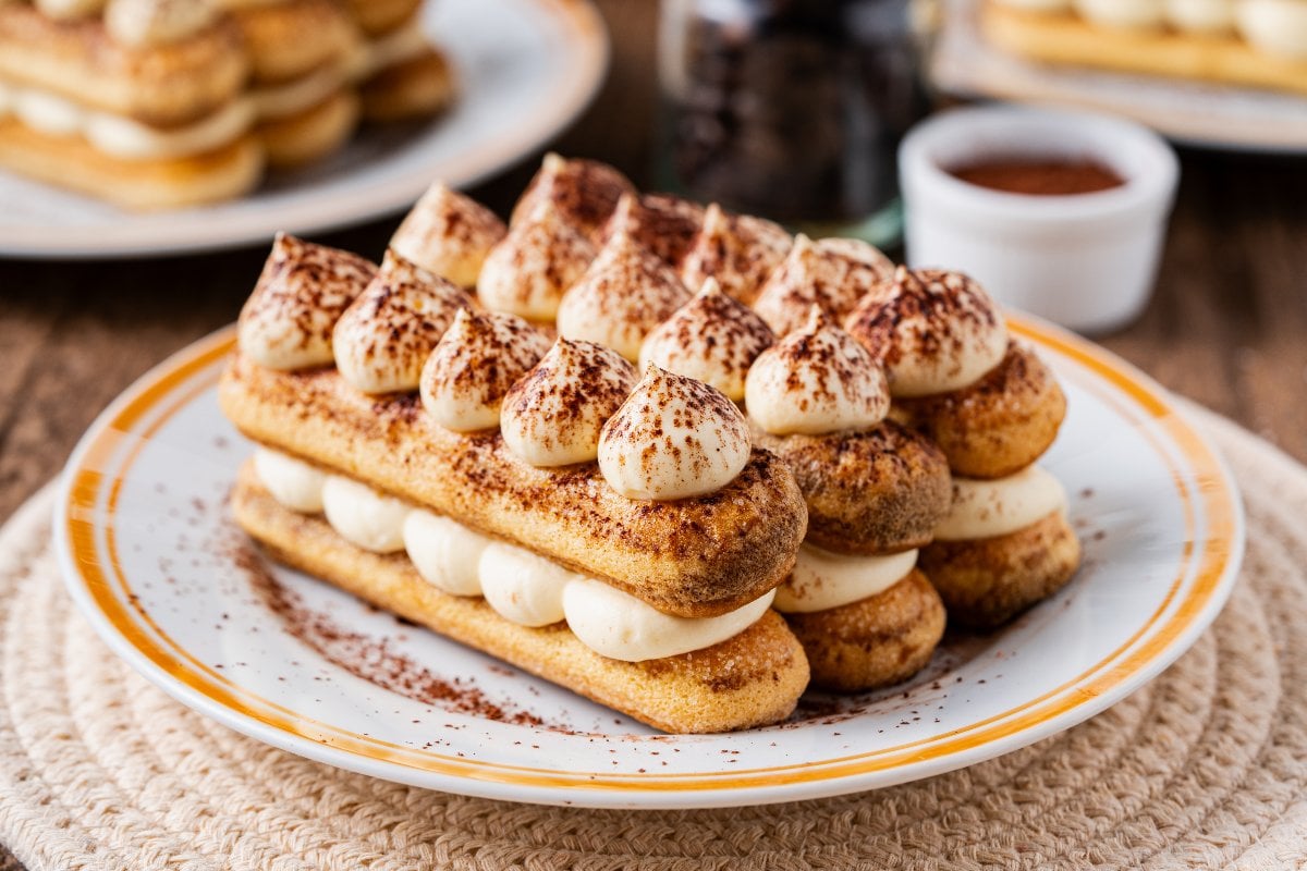 Three layers of individual tiramisu with piped cream and cocoa powder on a white plate, served on a woven placemat.