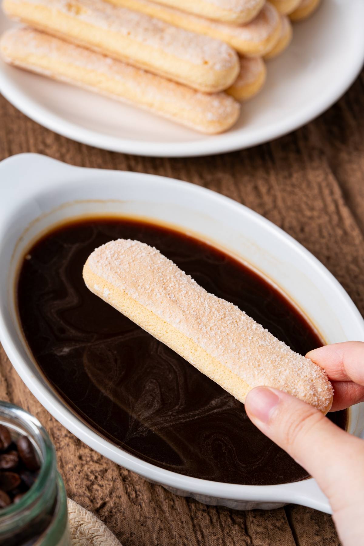 A hand dips a ladyfinger biscuit into a dish of dark coffee on a wooden table.