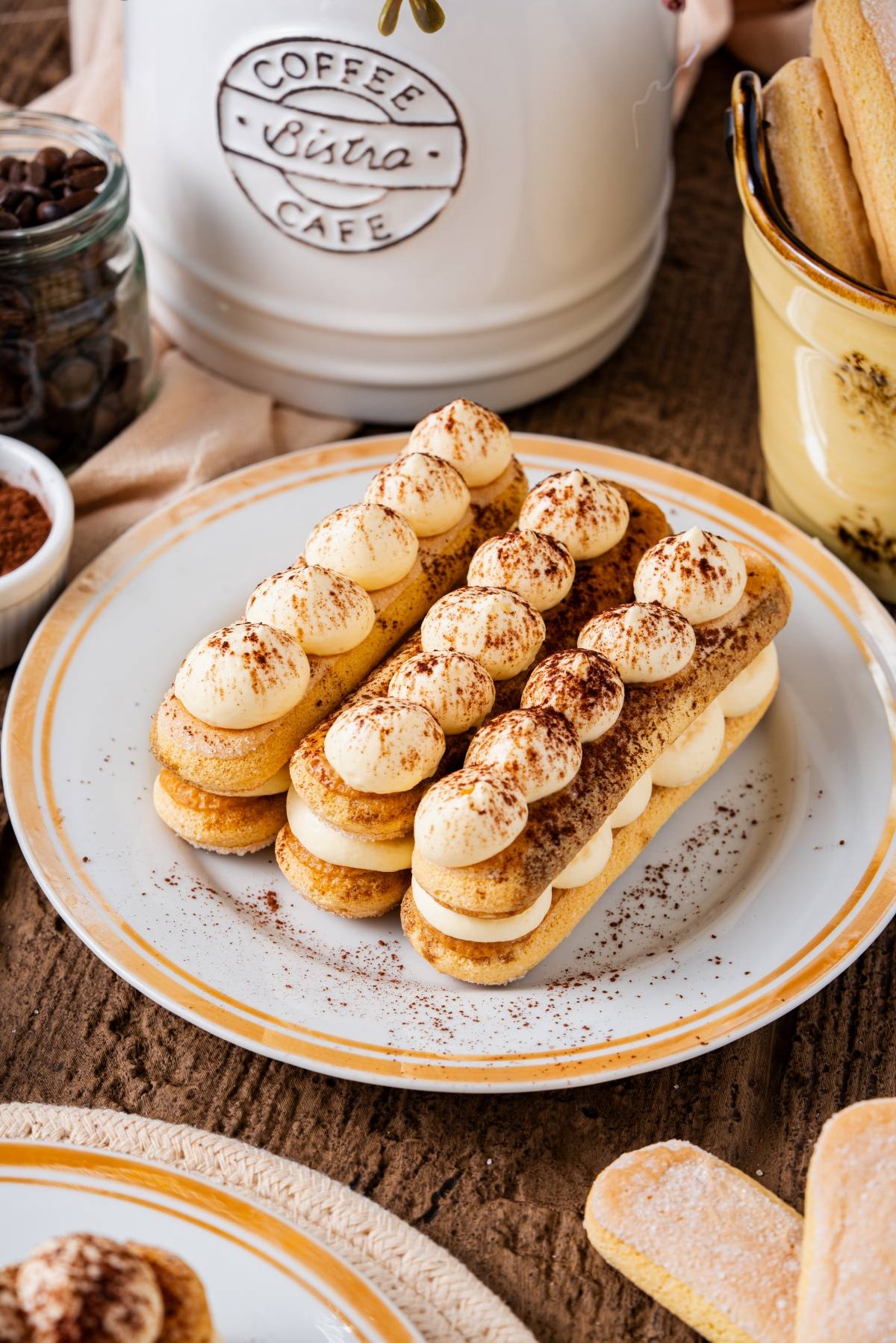 Tiramisu eclairs topped with cream and cocoa powder on a plate, with coffee-themed decor in the background.