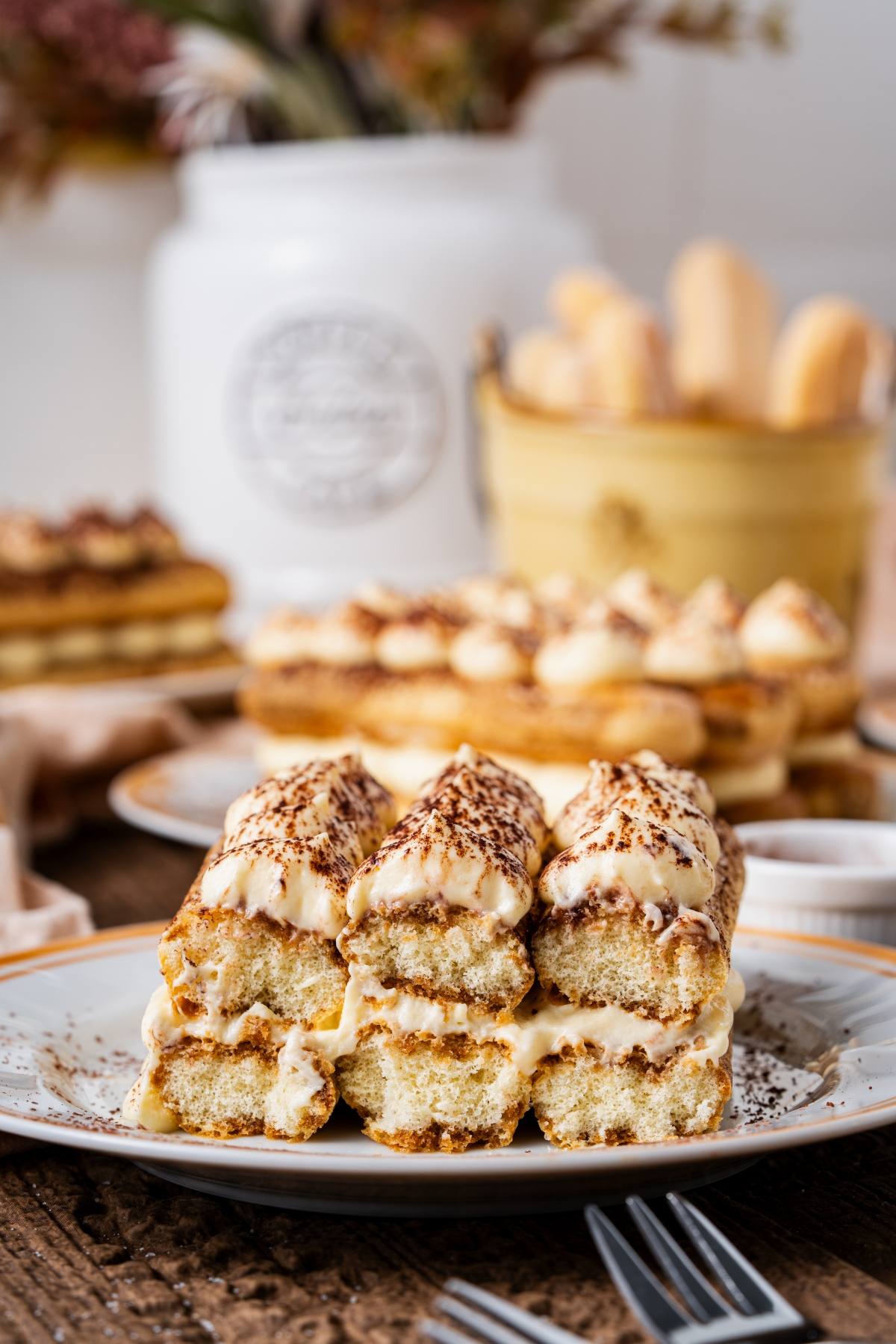 A close-up of tiramisu slices topped with cocoa powder, with more desserts and ladyfingers in the background.