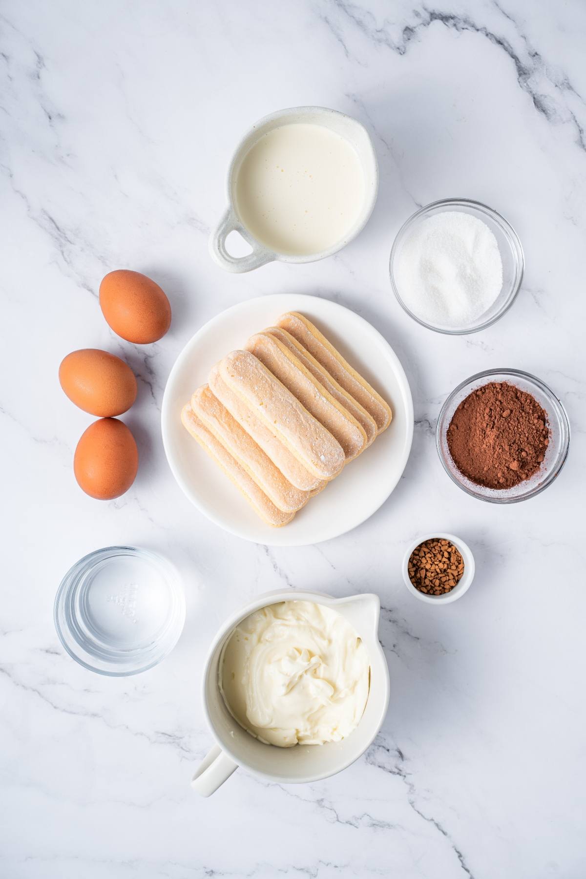 Tiramisu ingredients on a marble surface: eggs, ladyfingers, cream, sugar, cocoa powder, coffee, water, and mascarpone.
