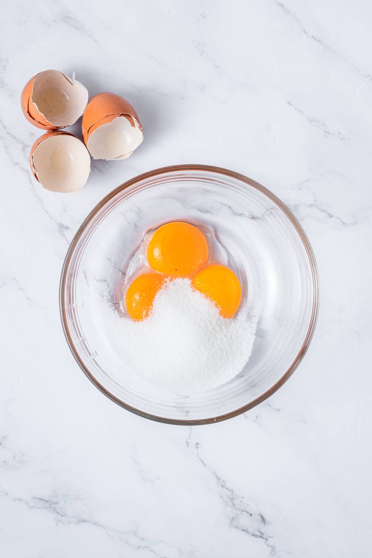 A glass bowl with three egg yolks and sugar, next to broken eggshells on a white marble surface.