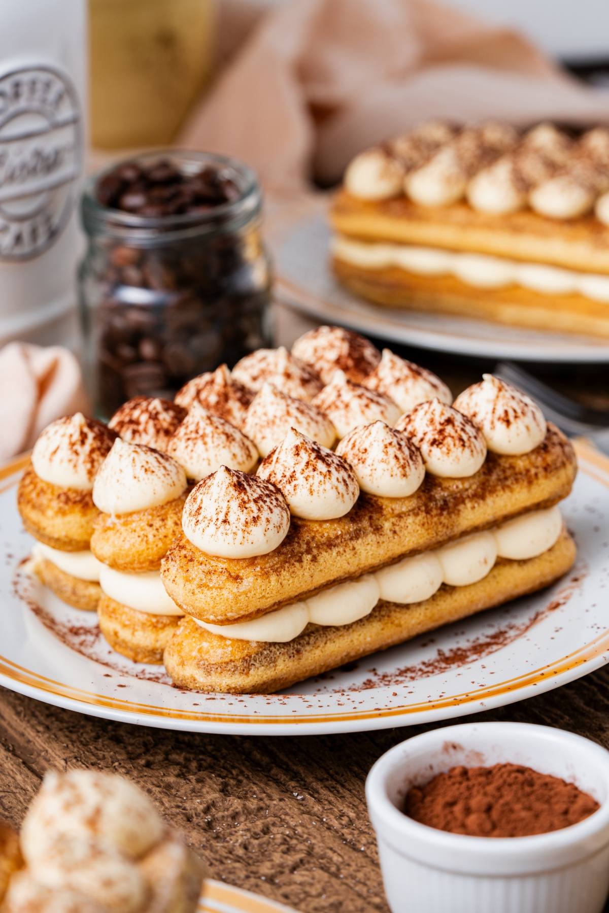 A plate of tiramisu with layers of cream and ladyfingers, dusted with cocoa powder, sits on a wooden table.