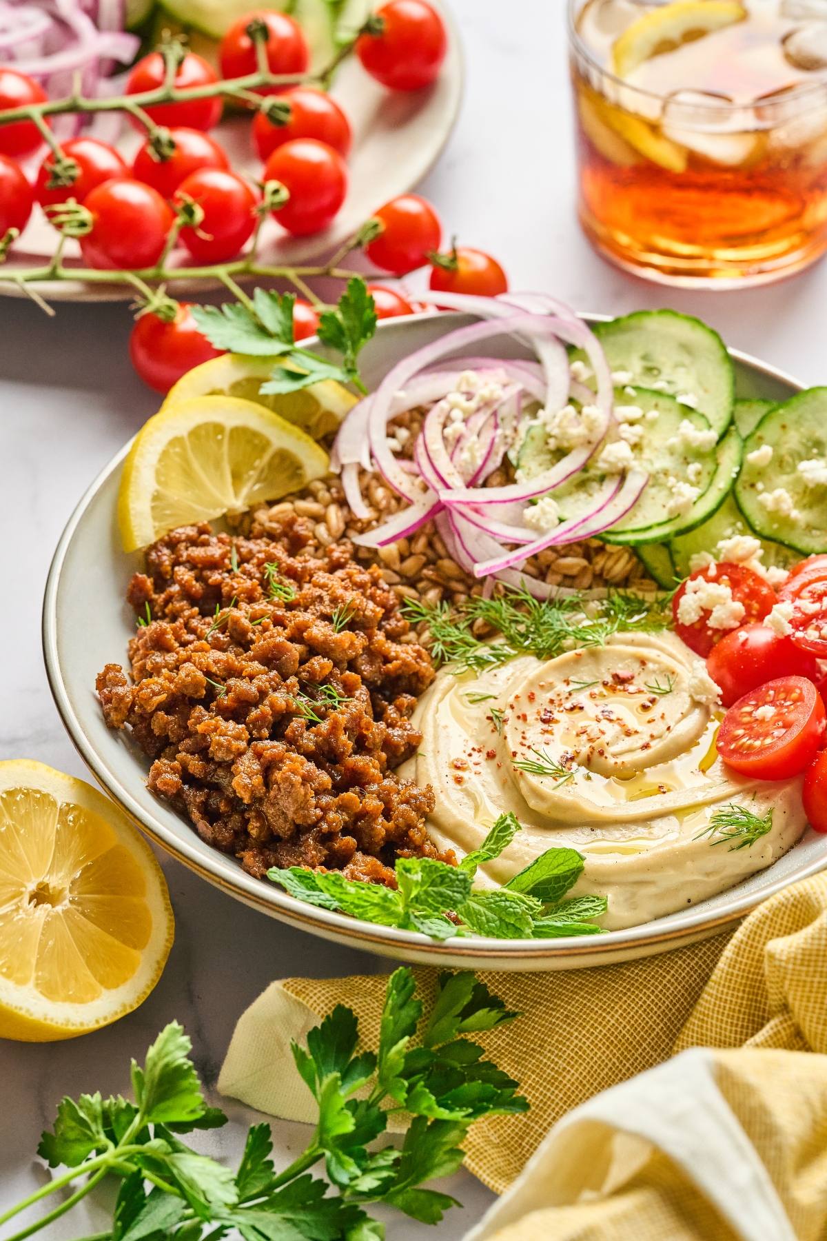 A bowl with hummus, ground meat, tomatoes, cucumbers, red onions, lemon, and herbs, next to a drink.
