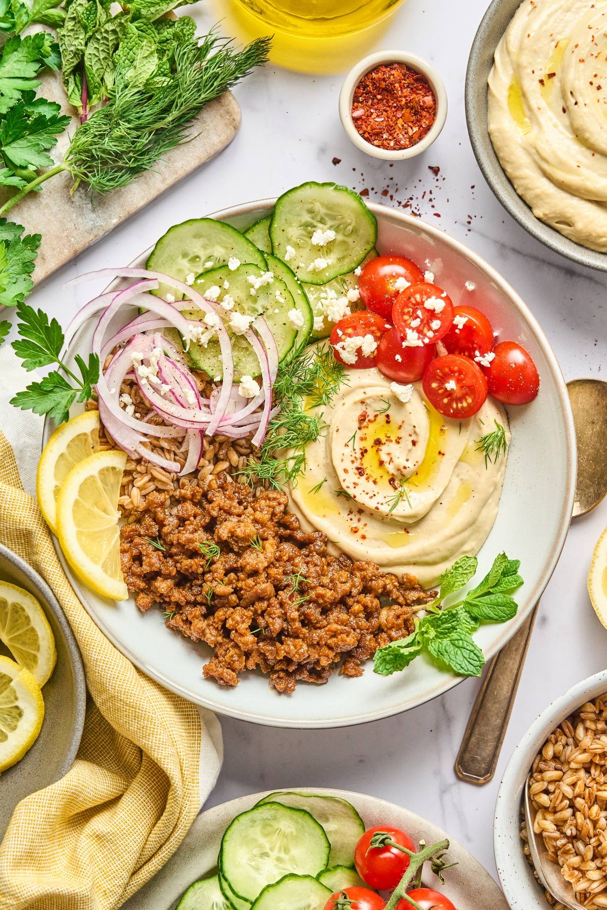 Bowl with hummus, spiced lentils, tomatoes, cucumber, red onion, herbs, and lemon slices on a white table.
