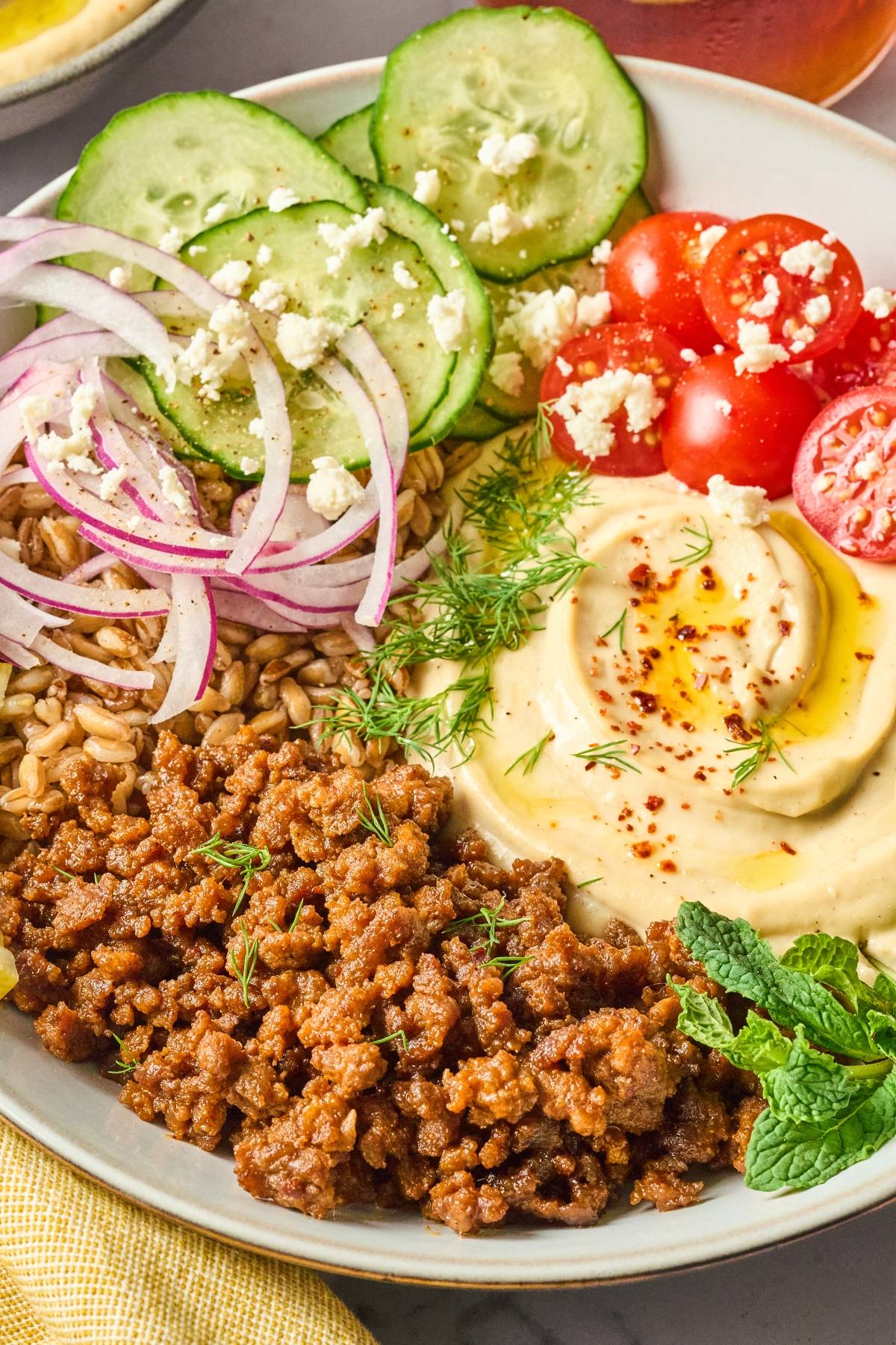 A bowl with plant-based ground meat, hummus, farro, cucumber, tomato, red onion, and fresh herbs.