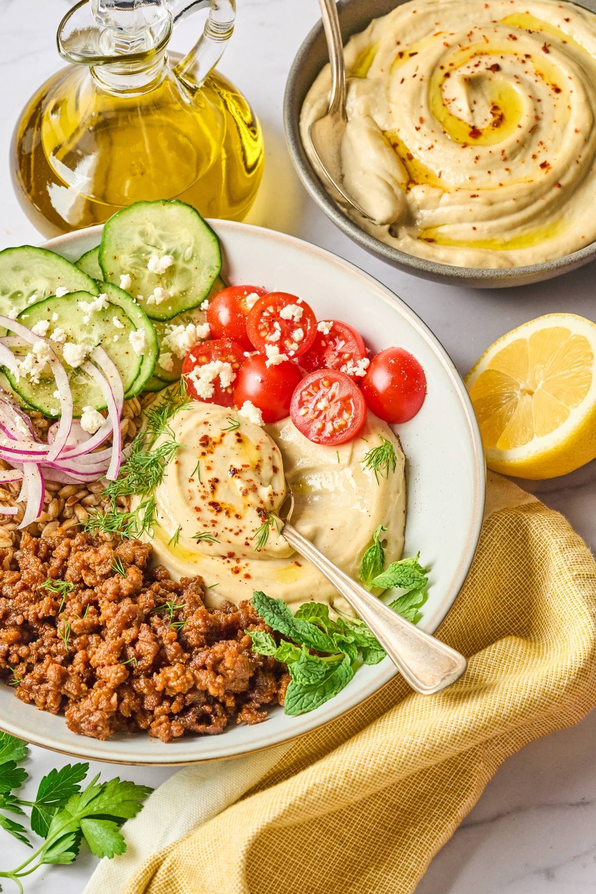 A bowl with hummus, ground meat, tomatoes, cucumbers, red onions, and herbs, next to olive oil and a lemon.