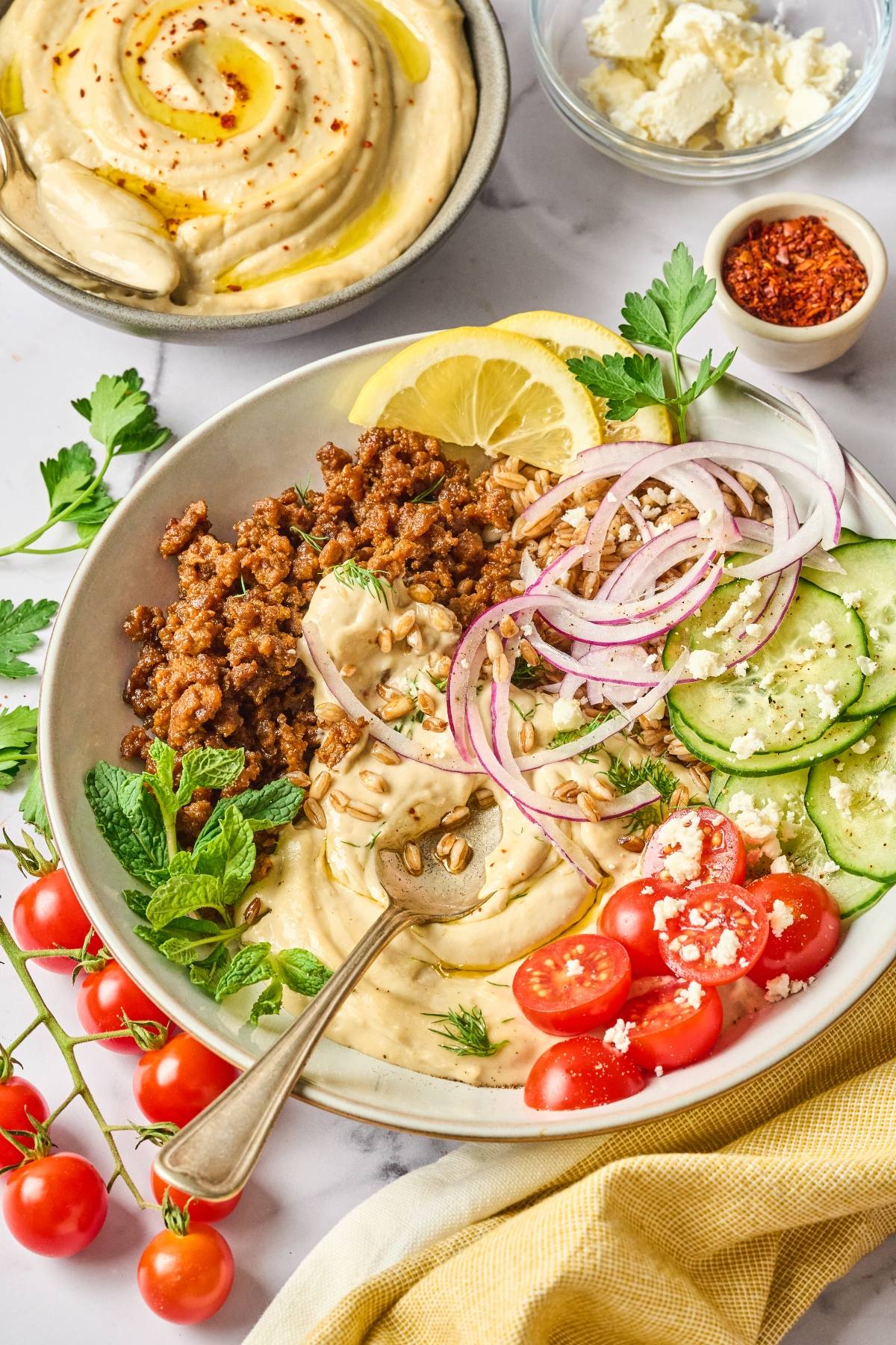 A bowl with hummus, ground meat, sliced cucumbers, tomatoes, onions, herbs, and lemon wedges on a table.