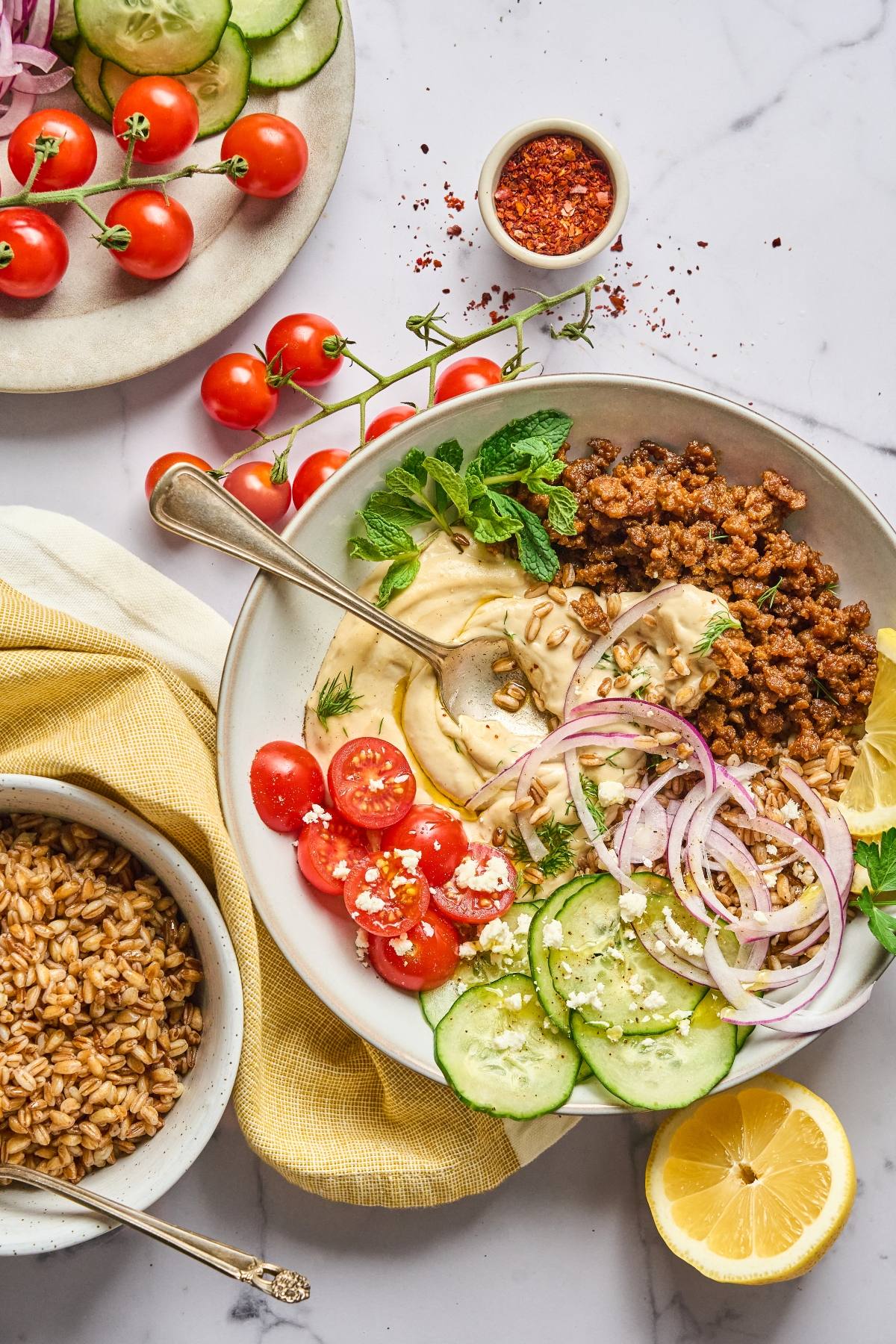 A bowl with ground meat, hummus, tomatoes, cucumber, red onion, feta, and herbs on a marble table.