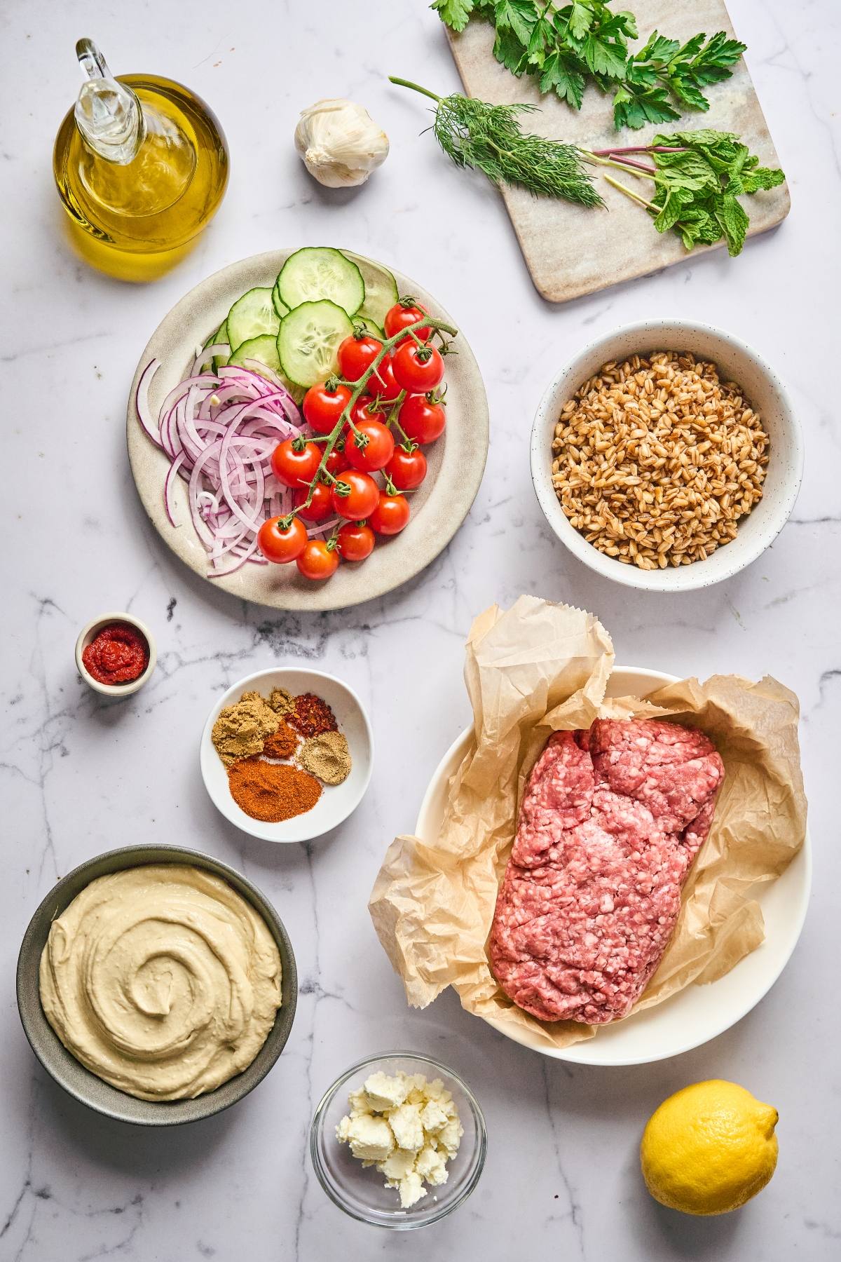 Overhead view of ground meat, spices, hummus, grains, vegetables, herbs, oil, and cheese on a marble surface.