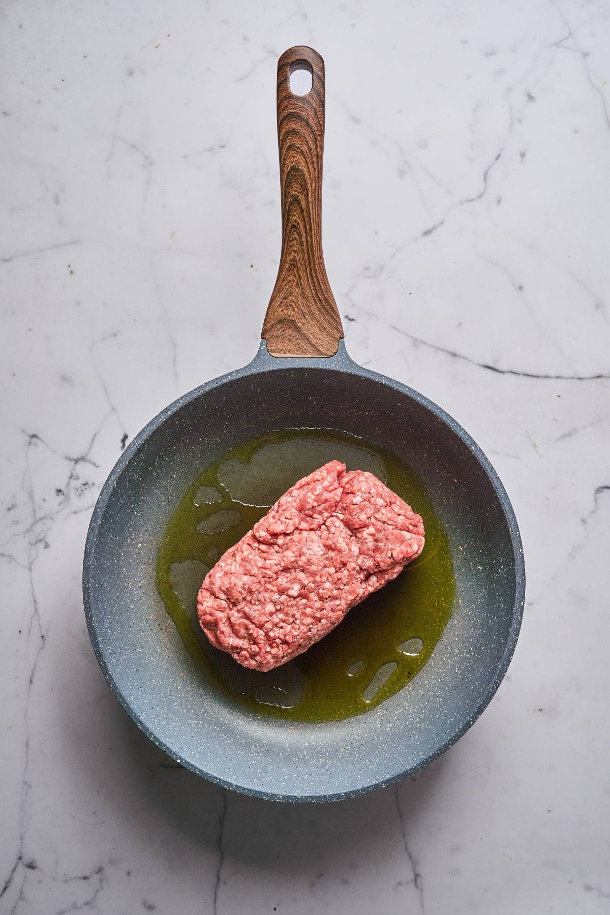 A raw block of ground meat in a frying pan with oil on a marble countertop.
