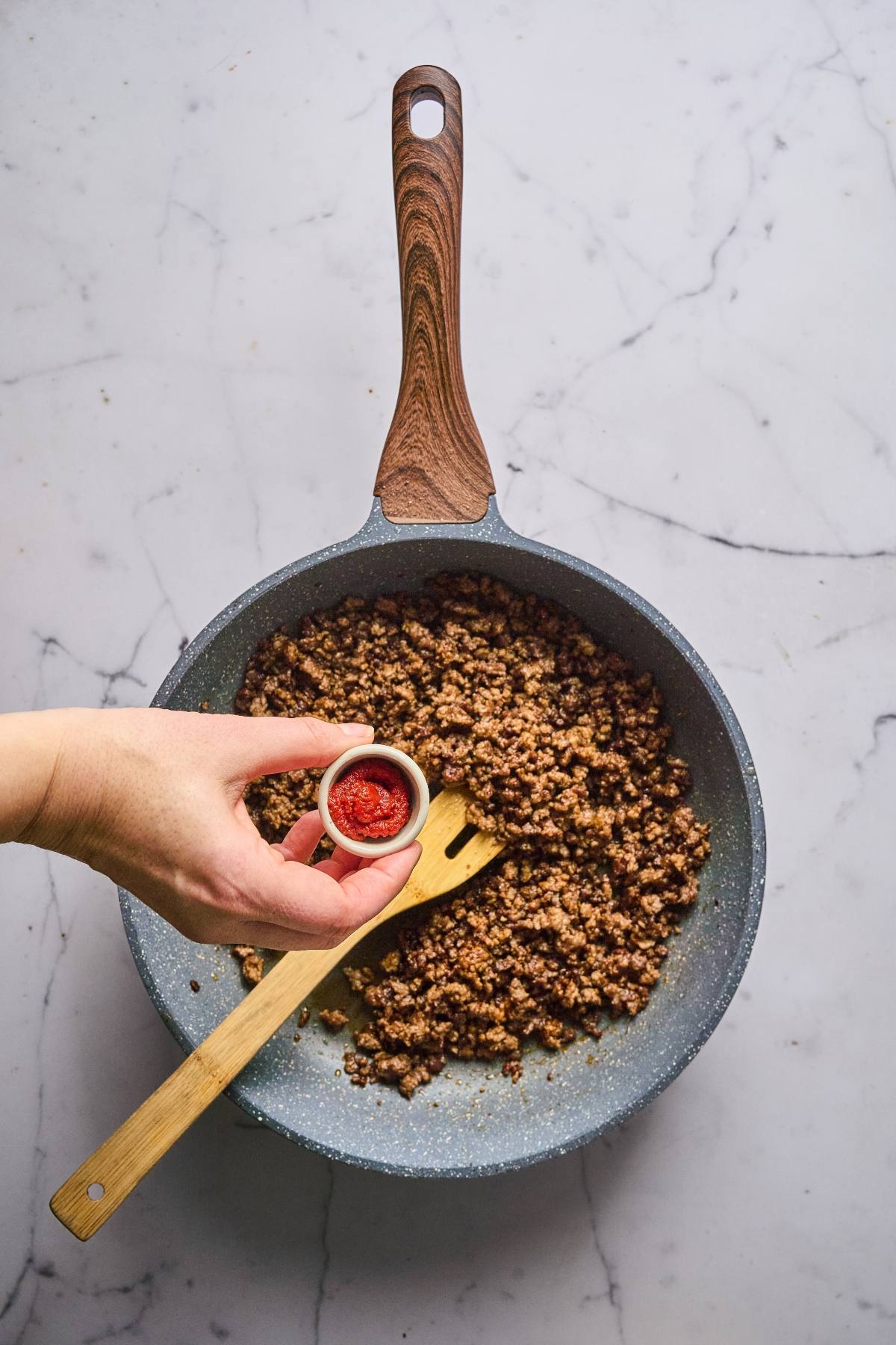 A hand adds red paste to cooked ground meat in a skillet with a wooden spatula on a marble counter.