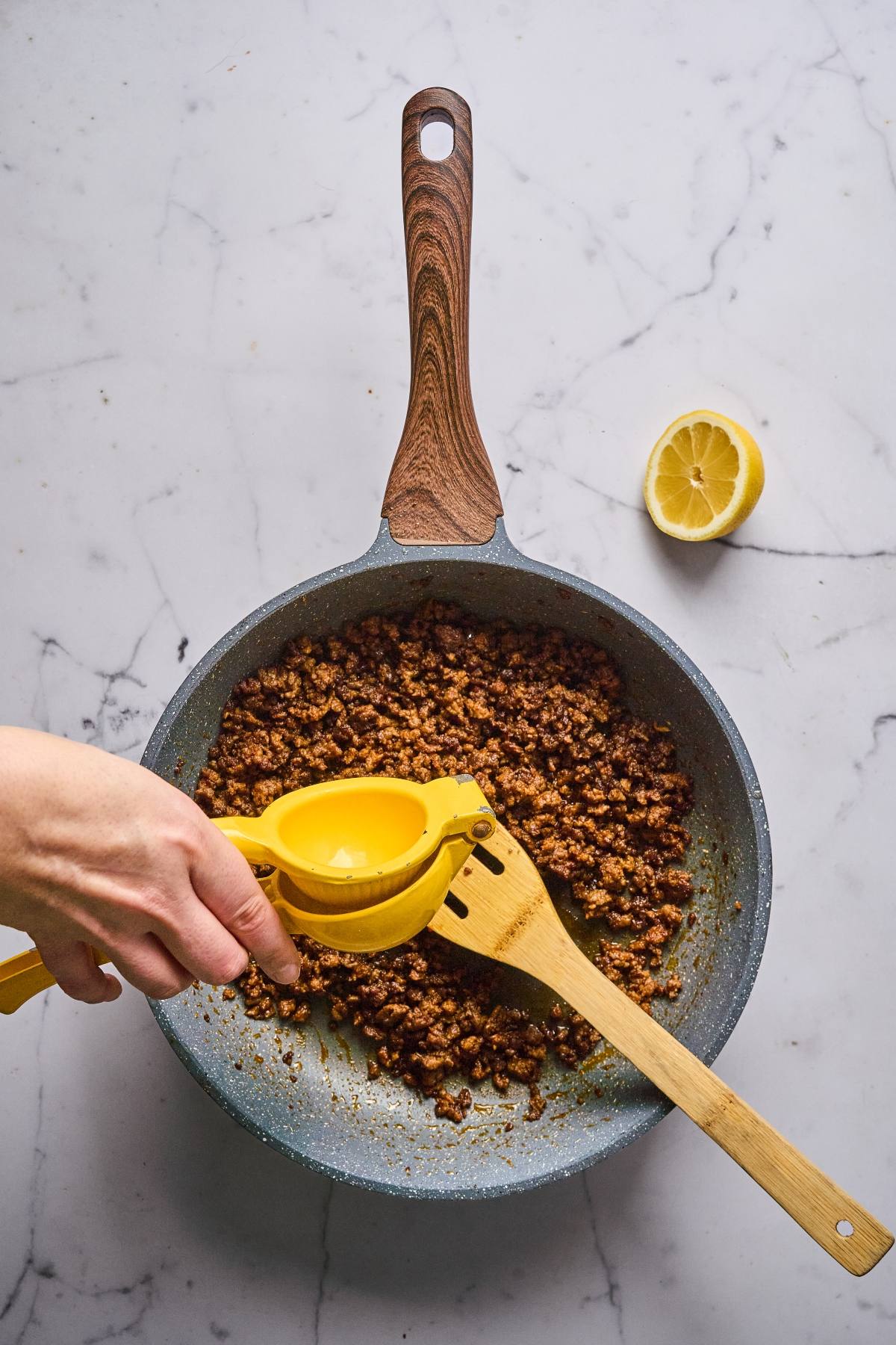 A hand squeezes lemon juice onto cooked ground meat in a skillet with a wooden spatula on a marble surface.