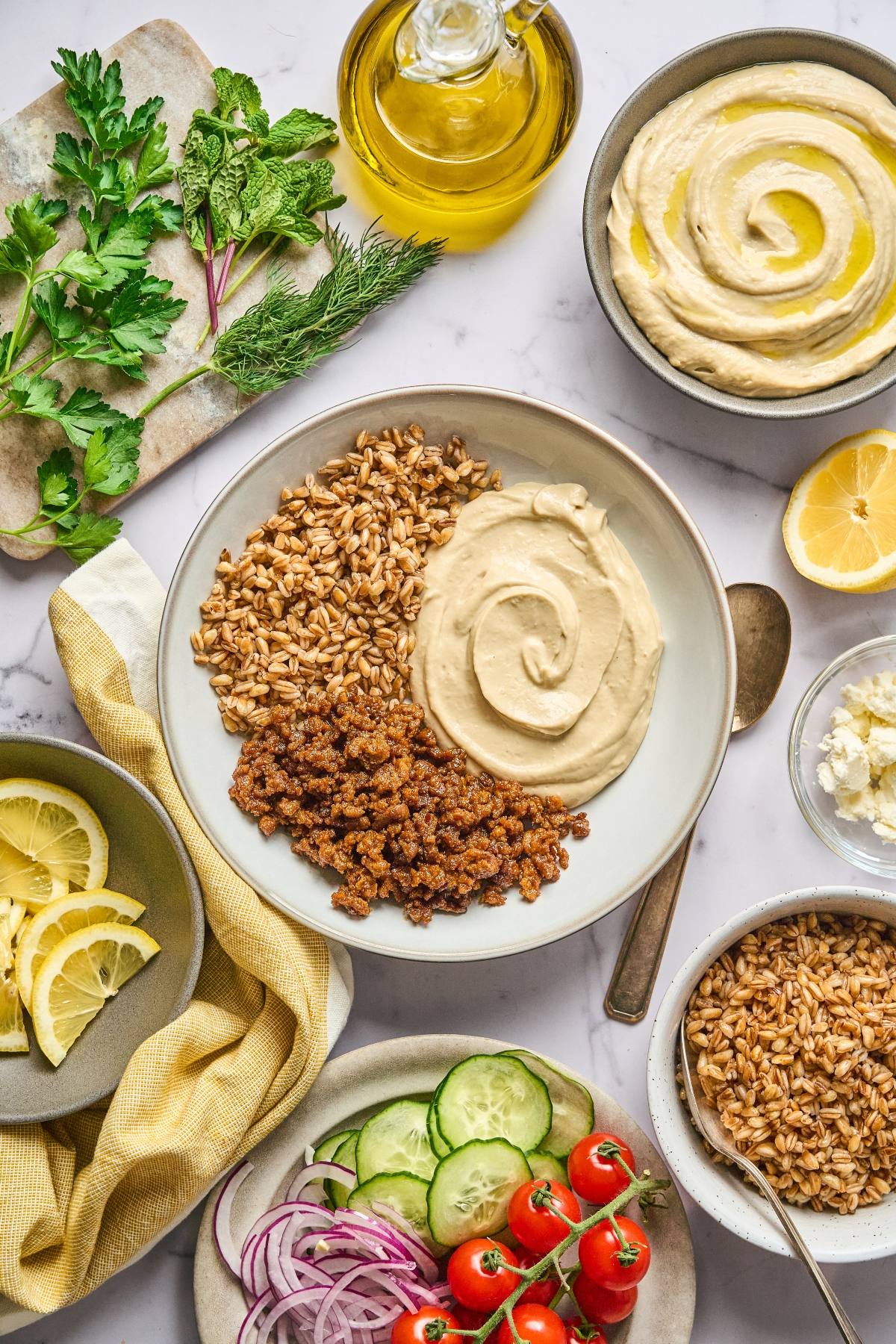 A bowl of hummus with grains and spiced crumbles, surrounded by herbs, veggies, lemon, and olive oil.