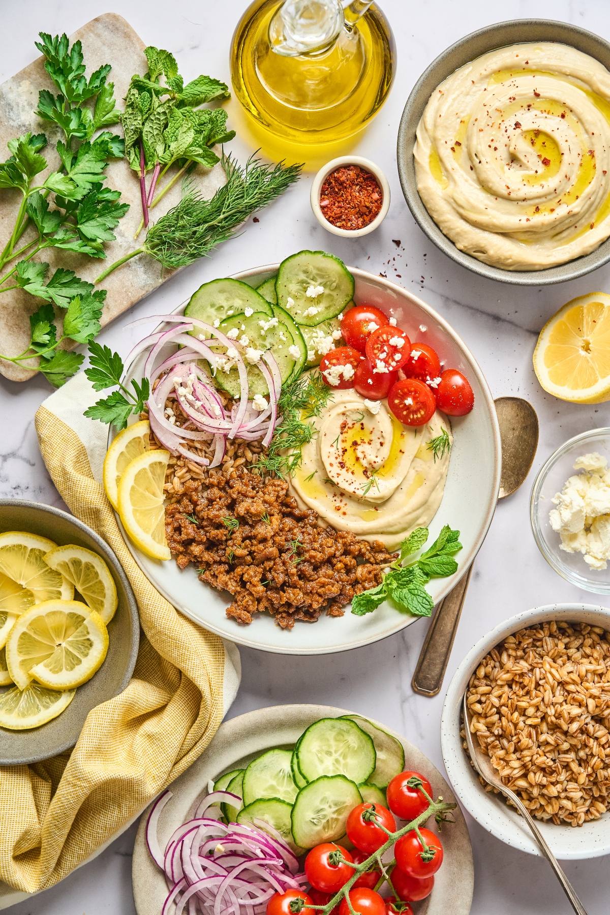 Bowl of hummus with lentils, cherry tomatoes, cucumber, onions, herbs, and lemon, surrounded by fresh ingredients.