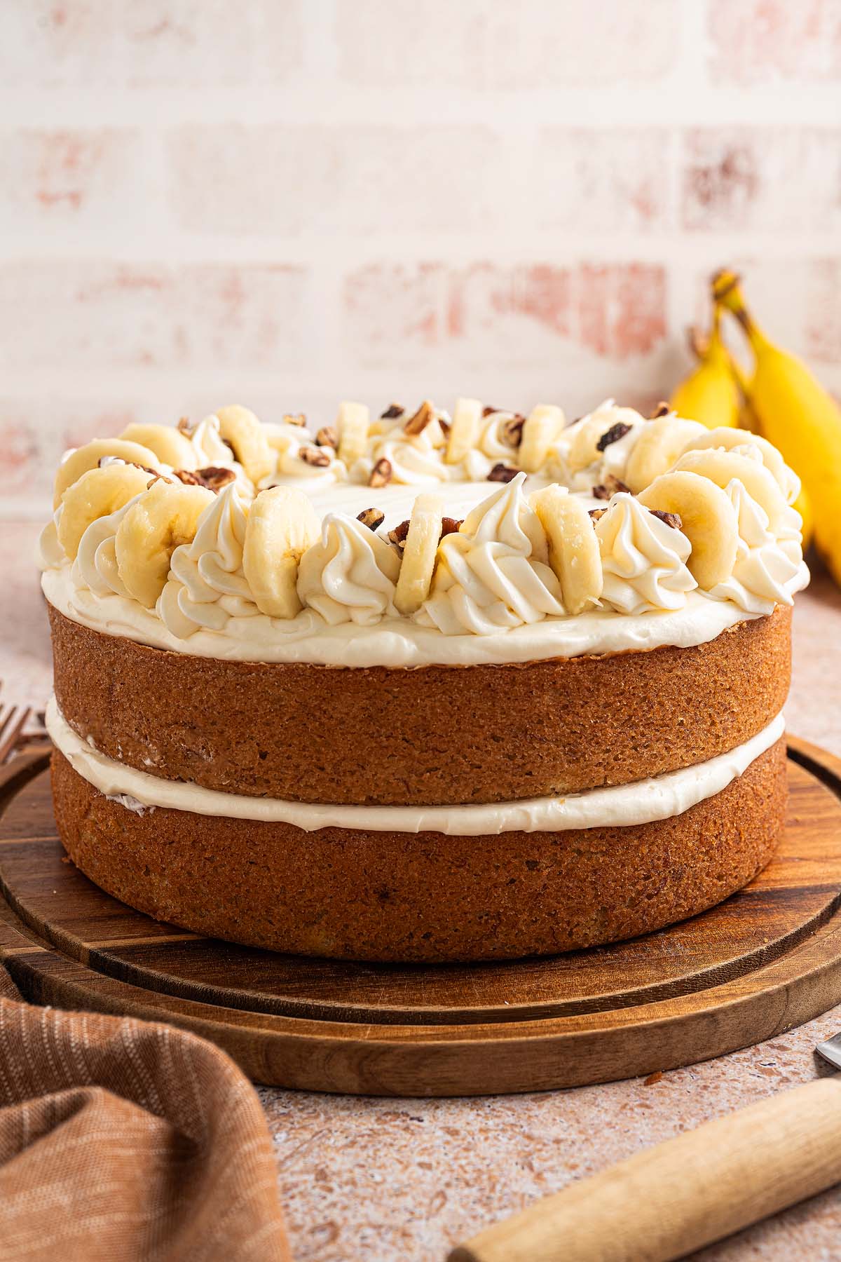 A delicious two-layer Banana Cake with white frosting and banana slices on top, beautifully displayed on a wooden board.
