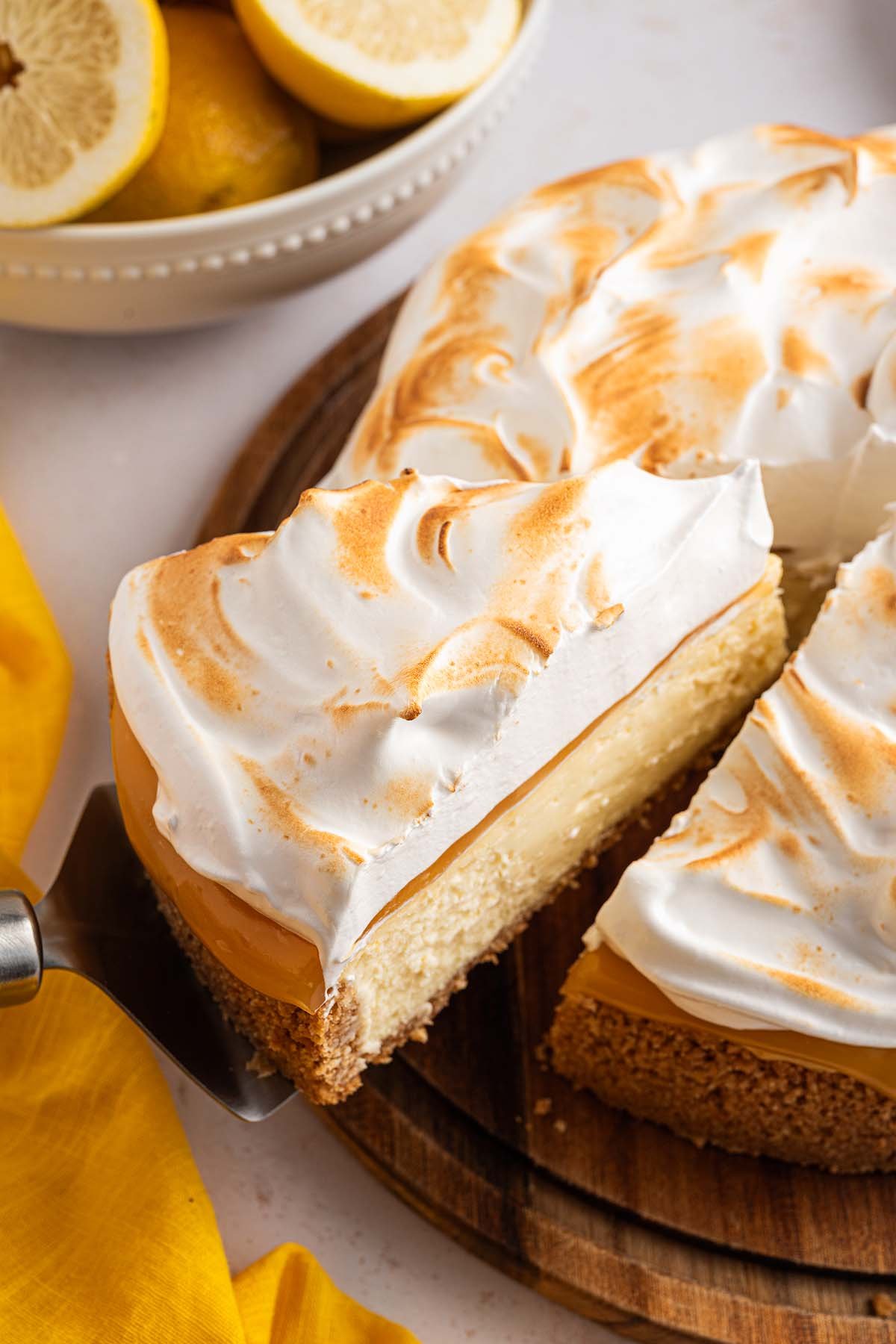 A slice of lemon meringue cheesecake being lifted from a whole pie, with fresh lemons in a bowl in the background.