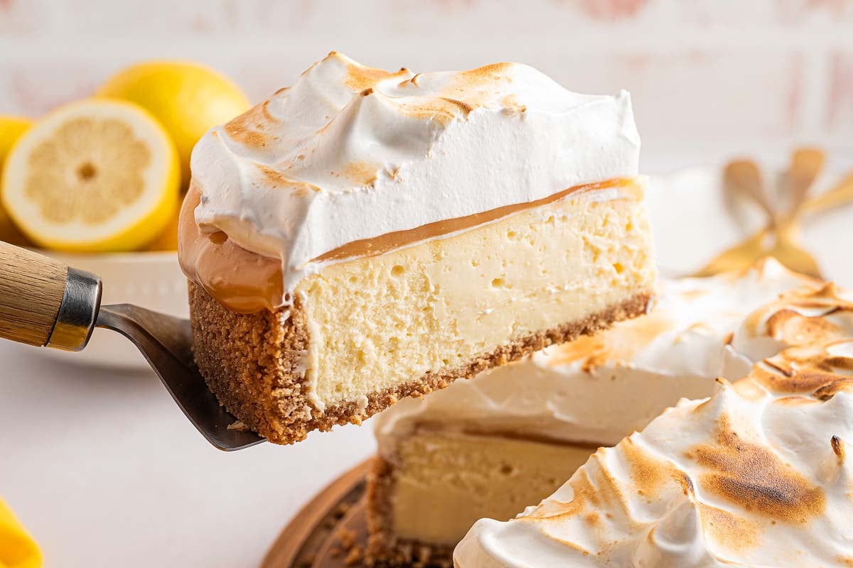 A slice of Lemon Meringue Cheesecake is being lifted, revealing creamy layers and a perfectly toasted meringue topping.