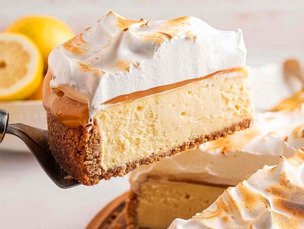 A slice of Lemon Meringue Cheesecake with caramel and fluffy meringue topping is being served from the whole cake.