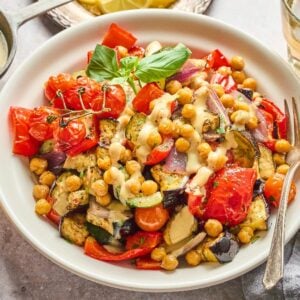 A Mediterranean Chickpea Skillet featuring roasted vegetables and chickpeas drizzled with sauce, served beside a glass of drink and a bowl of chickpeas.