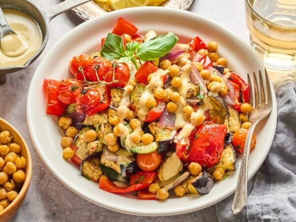 A Mediterranean Chickpea Skillet featuring roasted vegetables and chickpeas drizzled with sauce, served beside a glass of drink and a bowl of chickpeas.