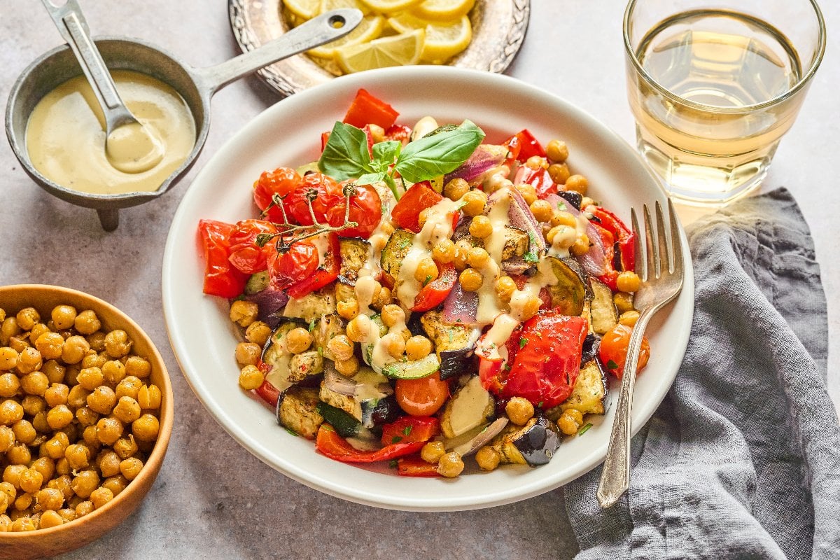 A Mediterranean Chickpea Skillet featuring roasted vegetables and chickpeas drizzled with sauce, served with a drink and extra chickpeas.