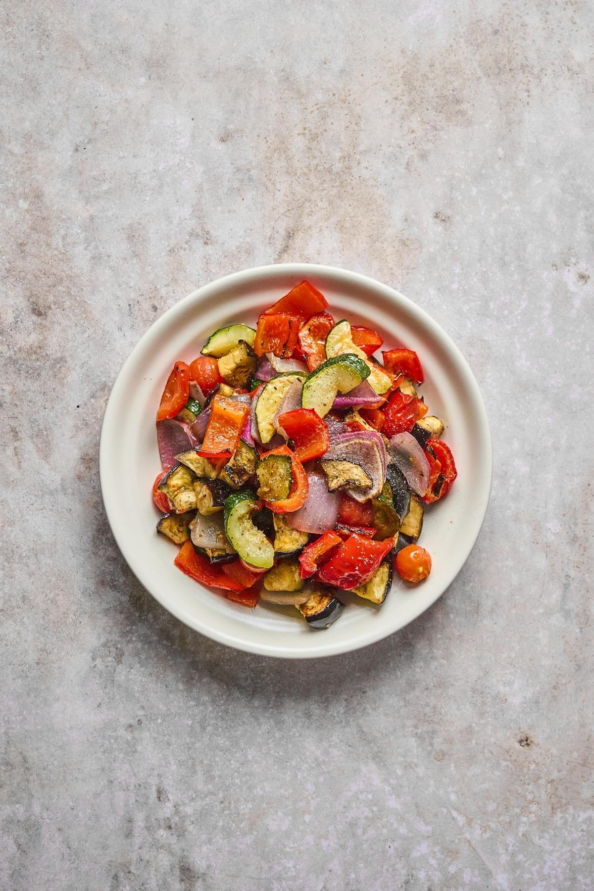 A white bowl filled with colorful roasted vegetables, including zucchini, peppers, onions, and tomatoes.