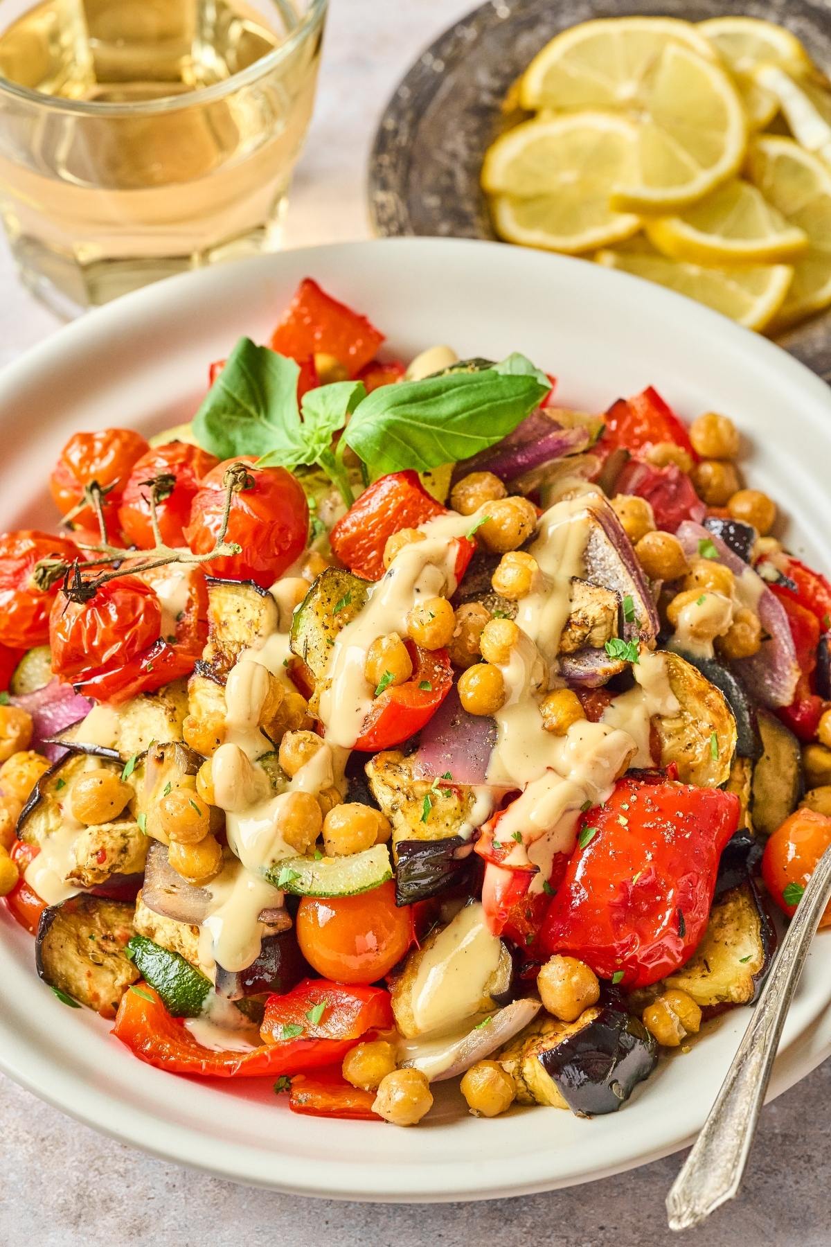A plate of roasted vegetables and chickpeas drizzled with sauce, garnished with basil, next to lemon slices.