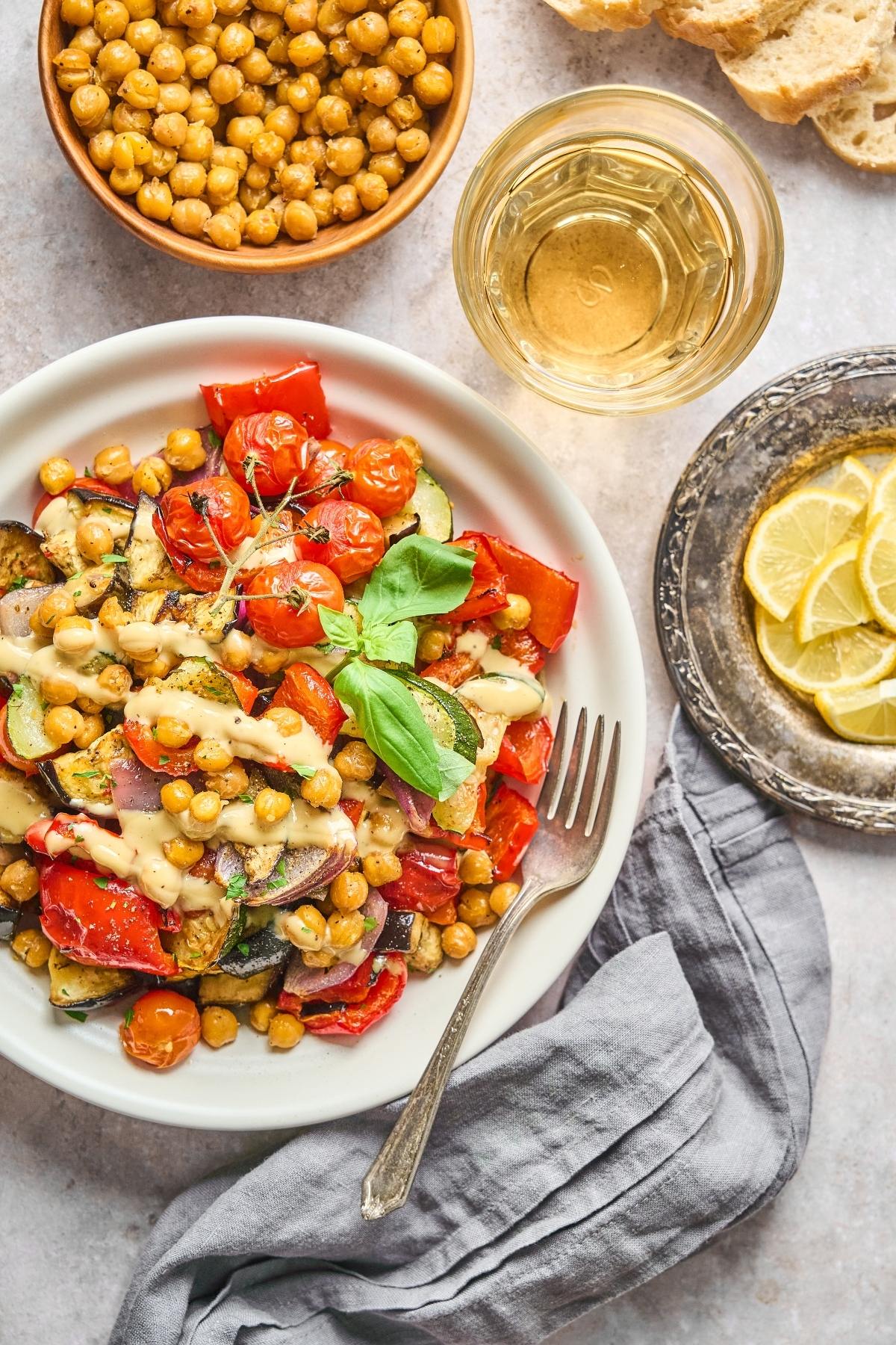 A plate of roasted vegetables and chickpeas with sauce, garnished with basil, next to lemon slices and a drink.