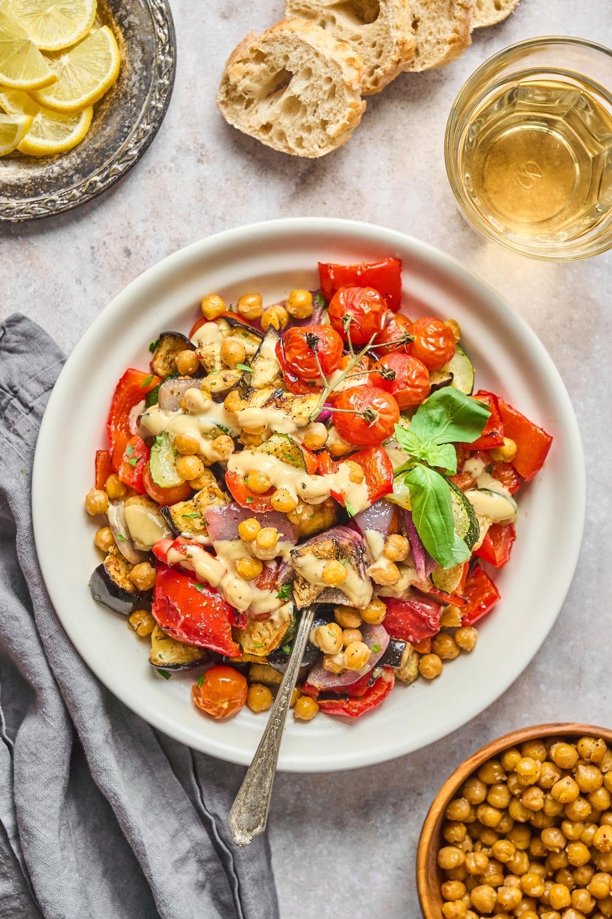 A bowl of roasted vegetables and chickpeas with fresh basil, next to bread, lemon slices, and a drink.