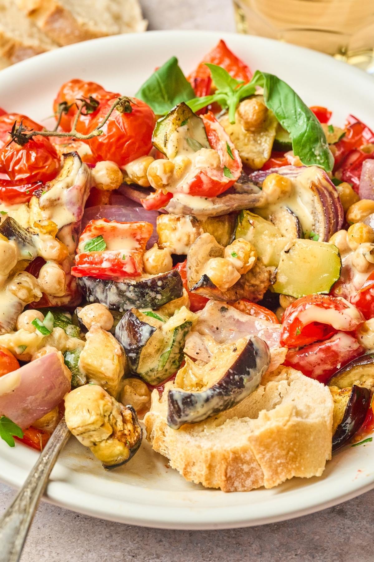 Colorful roasted vegetables and chickpeas salad with creamy dressing, served with a slice of bread on a white plate.