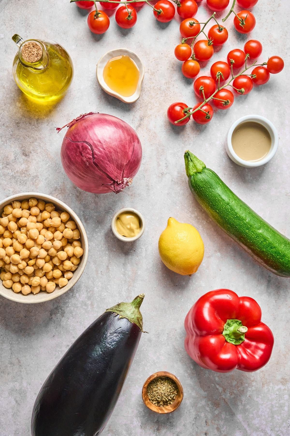 Colorful fresh vegetables, chickpeas, and condiments arranged on a light countertop, viewed from above.