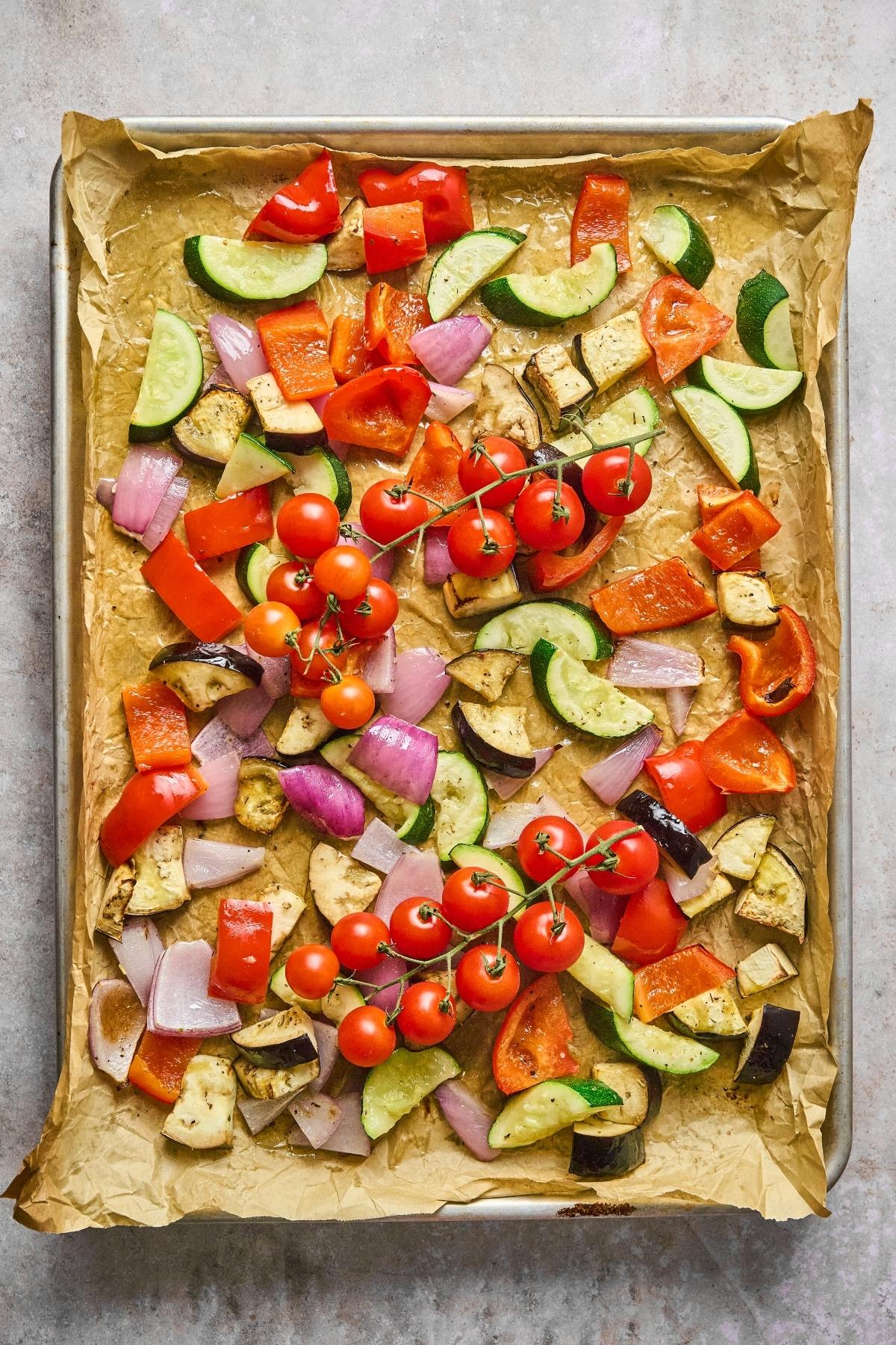 Colorful chopped vegetables and vine tomatoes on a parchment-lined baking sheet, ready to be roasted.