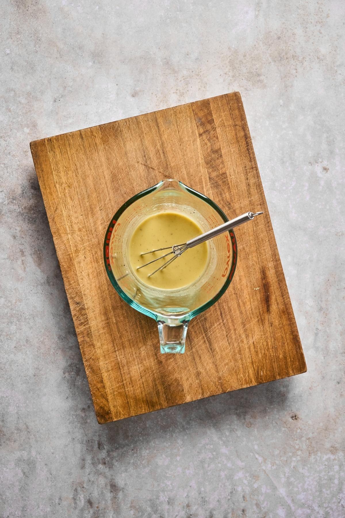 Glass measuring cup with creamy dressing and whisk on a wooden board, on a light gray surface.
