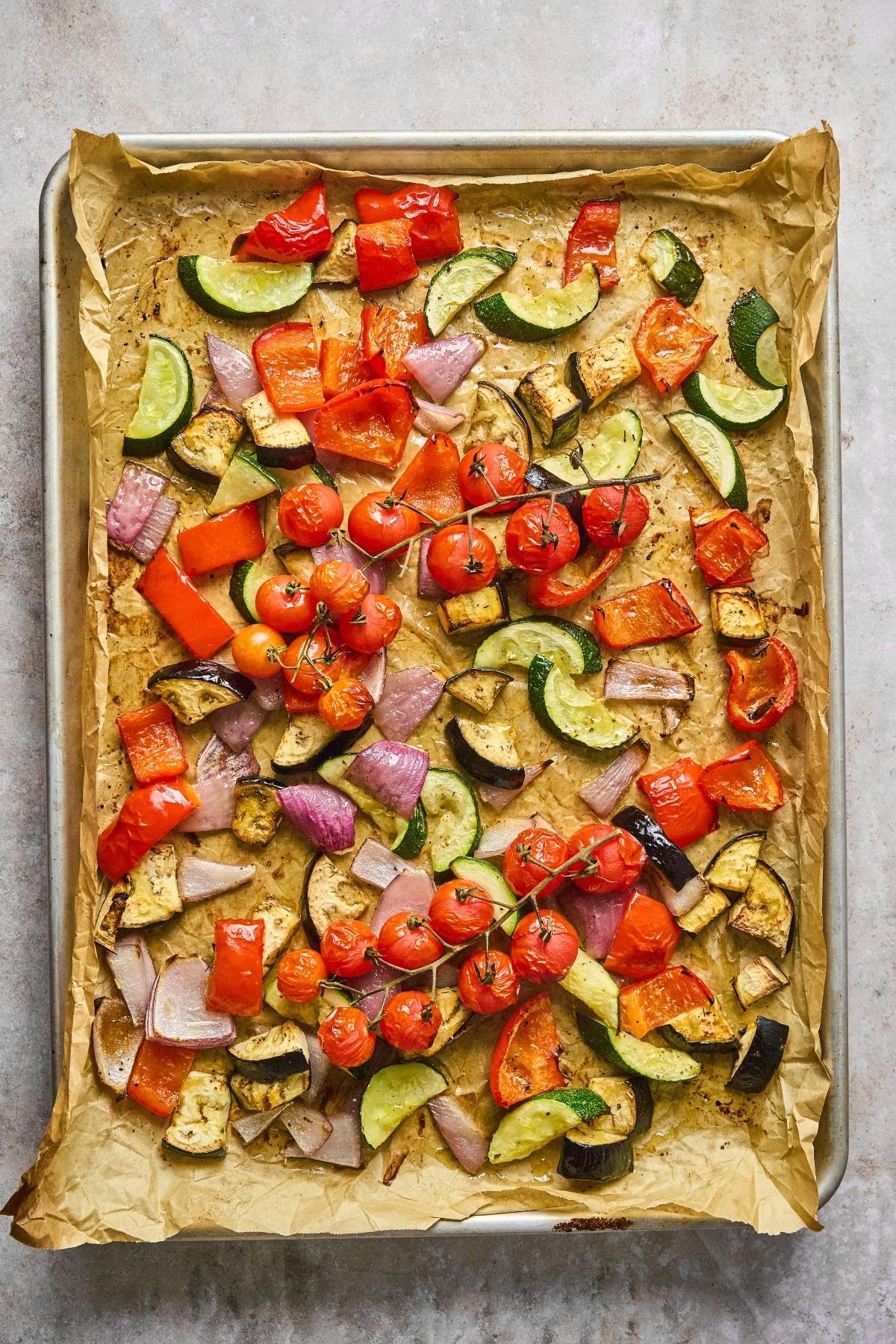Sheet pan of roasted vegetables including cherry tomatoes, zucchini, red peppers, onion, and eggplant on parchment paper.