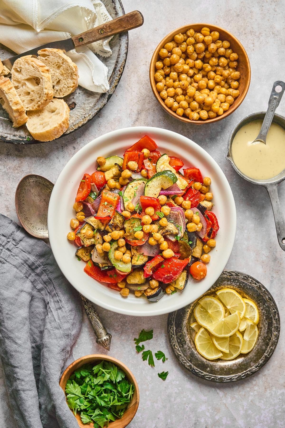 A bowl of roasted vegetables and chickpeas, surrounded by bread, lemon slices, sauce, and fresh herbs.