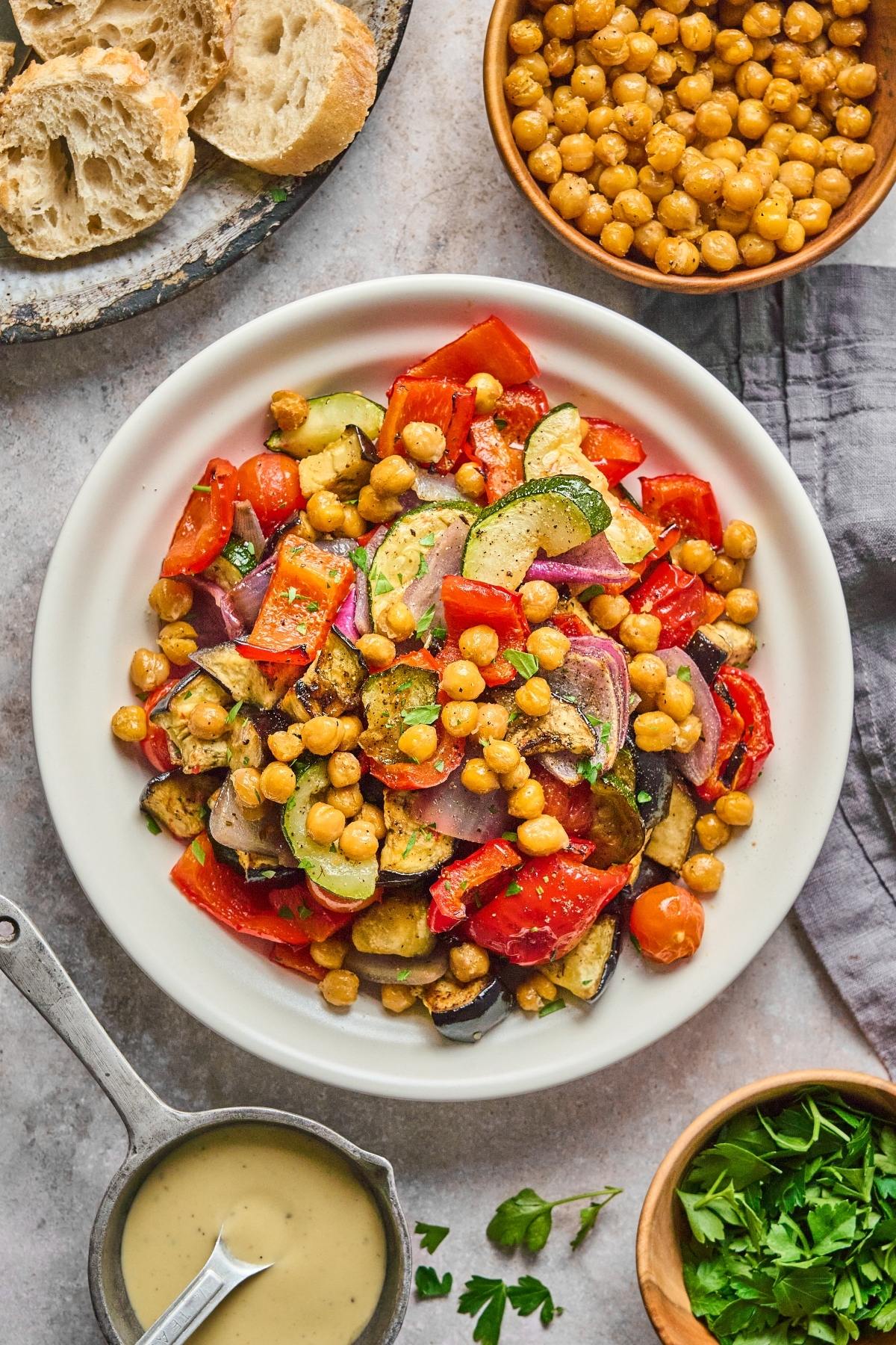 A plate of roasted vegetables and chickpeas, surrounded by bread, chickpeas, herbs, and a bowl of dressing.