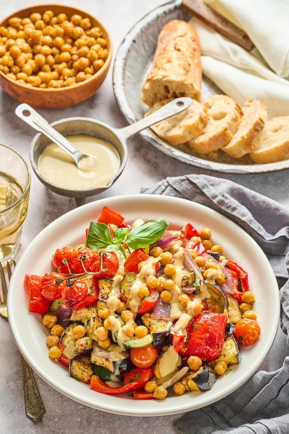 A bowl of roasted vegetables and chickpeas with tahini sauce, next to sliced bread and a bowl of chickpeas.